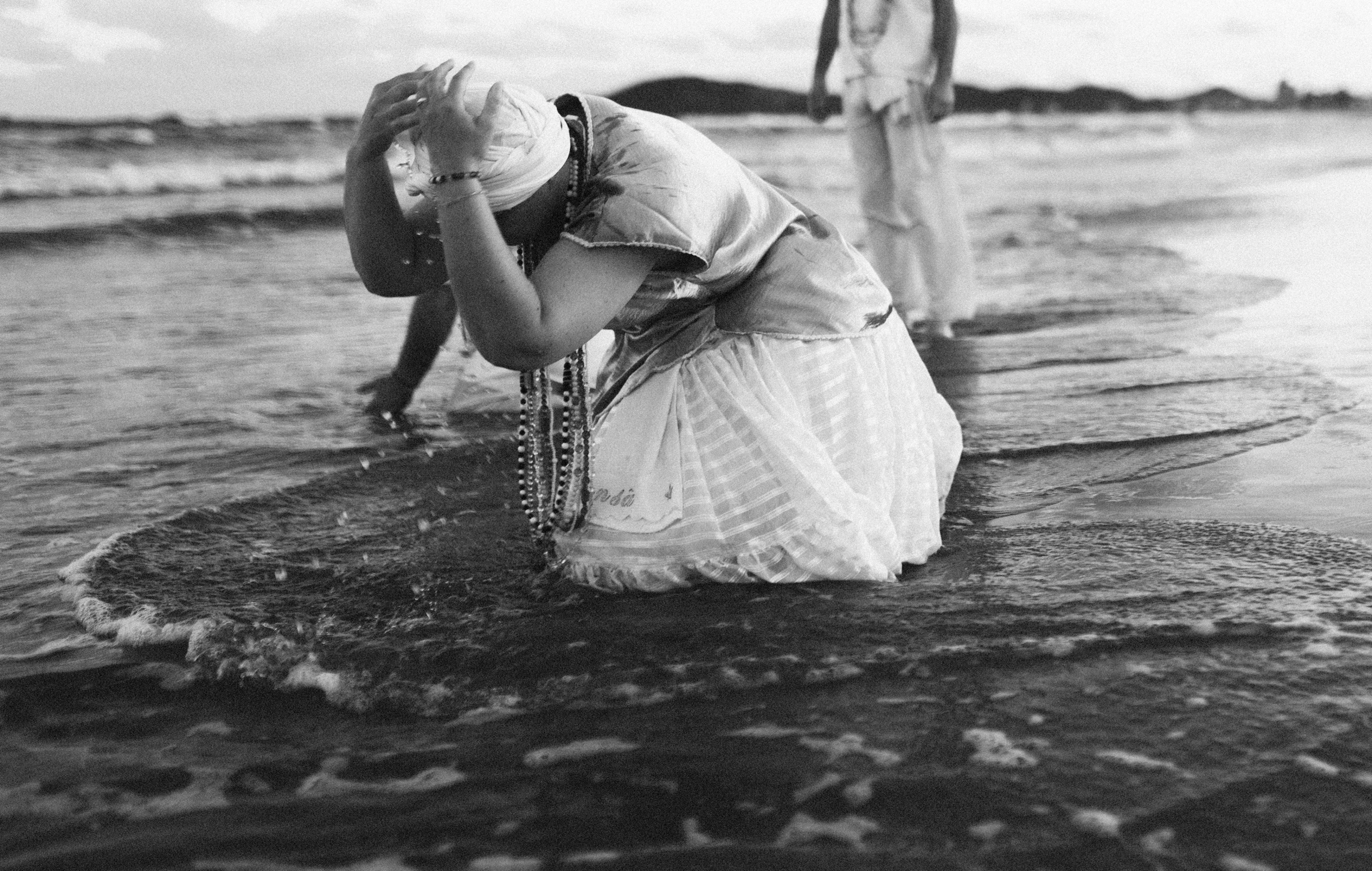 Audiodescrição da Imagem: Imagem em preto e branco. A fotografia mostra uma mulher ajoelhada à beira do mar, parcialmente submersa, realizando um ritual espiritual. Ela está com as mãos sobre a cabeça, como em um gesto de reverência ou oferenda, enquanto as ondas tocam suavemente seu corpo. A mulher veste roupas típicas de umbanda: uma saia rodada branca com detalhes rendados, uma blusa brilhante de tecido leve e um turbante branco na cabeça. Colares de contas coloridas pendem do seu pescoço, simbolizando sua ligação com os orixás. Outras pessoas também aparecem ao fundo, parcialmente visíveis, e usam trajes semelhantes. A maré está baixa, e o cenário ao redor inclui o mar calmo e uma linha de montanhas escuras no horizonte, sob um céu nublado.