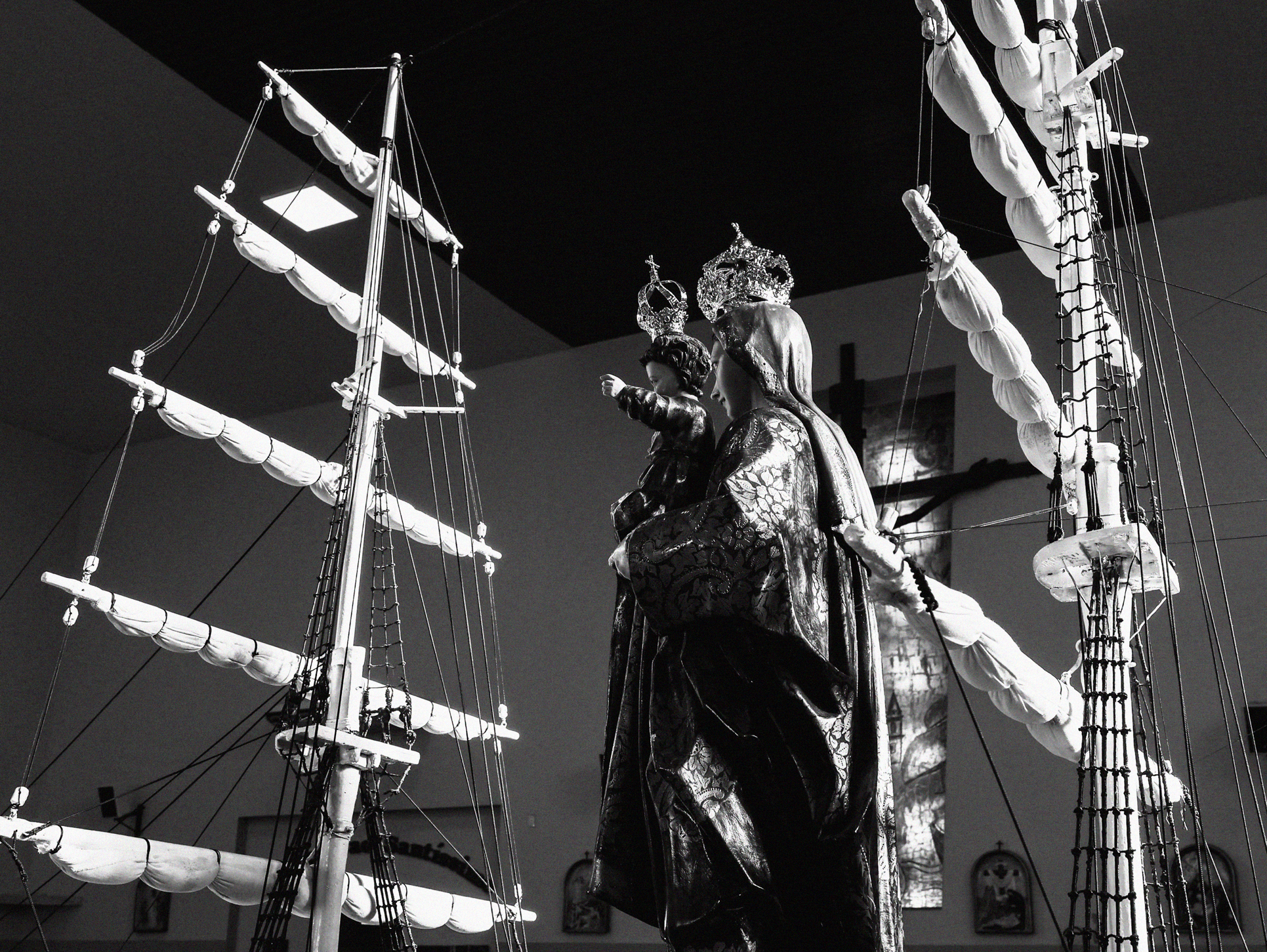 Imagem 3 da Galeria de Fotos. Audiodescrição: Fotografia em preto e branco, capturada em ambiente interno, possivelmente o interior de uma igreja. Em destaque, no centro da imagem, está uma escultura de Nossa Senhora dos Navegantes com o Menino Jesus nos braços, ambos usando mantos detalhados e coroas ornamentadas. A figura sagrada está posicionada entre duas grandes estruturas de velame, que simulam mastros e velas de um navio — remetendo ao tema marítimo, típico da iconografia de devoção à padroeira dos pescadores e navegantes. As velas estão enroladas e dispostas horizontalmente em várias fileiras ao longo dos mastros, com cordas, redes e elementos náuticos cuidadosamente reproduzidos. Os mastros, à esquerda e à direita da escultura, criam uma moldura simbólica que reforça a ligação entre a fé e o mar. Ao fundo, é possível ver vitrais parcialmente iluminados, um crucifixo pendurado na parede e alguns quadros religiosos decorando o espaço. A iluminação incide lateralmente, destacando os detalhes do tecido brocado do manto de Nossa Senhora, o brilho das coroas e os volumes das velas e cordas, criando uma atmosfera de reverência, solenidade e espiritualidade. A composição sugere uma instalação artística ou altar devocional voltado à proteção divina sobre os navegantes, expressando fé, tradição e simbolismo marinho.