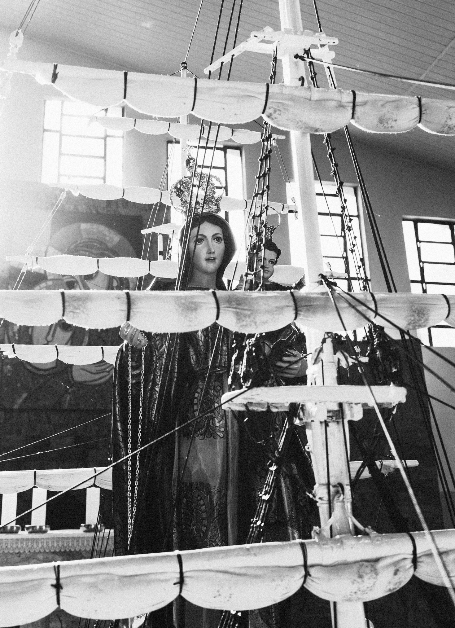 Imagem 4 da Galeria de Fotos. Audiodescrição: Fotografia em preto e branco, capturada em ambiente interno com iluminação natural intensa, mostrando uma imagem religiosa de Nossa Senhora dos Navegantes com o Menino Jesus nos braços, parcialmente encoberta por uma estrutura simbólica de embarcação, composta por mastros altos, cordames e velas enroladas em níveis horizontais. A imagem está inserida entre os mastros, de forma que os elementos náuticos parecem integrar o altar. Nossa Senhora aparece com olhar sereno e direcionado à frente, usando um manto escuro com desenhos ornamentais e uma coroa sobre a cabeça. O Menino Jesus também usa uma pequena coroa e está posicionado no braço esquerdo da santa. A imagem transmite delicadeza e solenidade. As velas, fixadas em vários níveis dos mastros, formam um padrão visual que atravessa a composição horizontalmente, criando uma espécie de “gaiola” simbólica em torno da figura sagrada. Corda, correntes e cordames reforçam o tema marítimo. Ao fundo, vê-se parte do interior de uma igreja: paredes claras, janelas retangulares, altar e objetos litúrgicos desfocados. A iluminação vinda da esquerda — provavelmente luz solar entrando pelas janelas — banha a cena e cria reflexos sobre os tecidos e coroas, ampliando a atmosfera sagrada e devocional. A composição enfatiza a devoção marinha, em que a santa é invocada como protetora dos navegantes e pescadores.