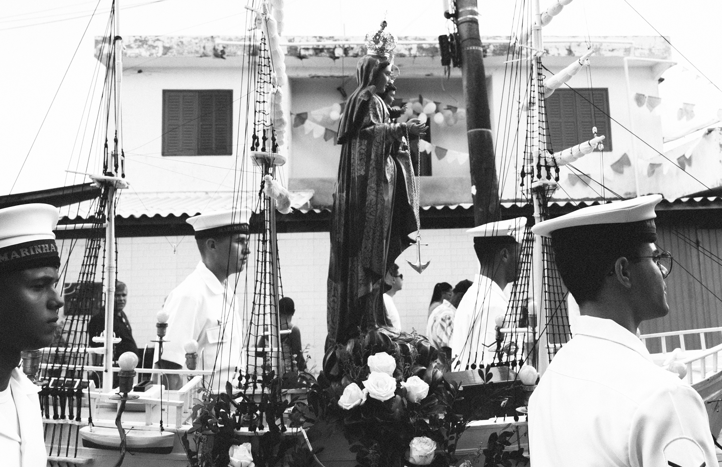 Imagem 5 da Galeria de Fotos. Audiodescrição: Fotografia em preto e branco, capturada em ambiente externo, durante o dia, retratando uma procissão religiosa. No centro da imagem, a escultura de Nossa Senhora dos Navegantes está posicionada sobre uma plataforma decorada em formato de embarcação. A santa está vestida com manto ornamentado e coroa, segurando um escapulário com uma pequena âncora pendente, símbolo de proteção aos navegantes. Ao seu lado, o Menino Jesus também aparece em menor escala, em postura abençoando. A embarcação simbólica tem mastros com velas enroladas e cordames, simulando um navio à vela, cercado por flores brancas, principalmente rosas, dispostas na base da imagem. A procissão é conduzida por quatro homens vestidos com uniforme branco da Marinha Brasileira, com quepes e postura formal, ladeando a embarcação devocional. No chapéu de cada um é possível ler a palavra “MARINHA”, indicando a participação institucional no cortejo. Ao fundo, há um prédio de dois andares com janelas fechadas e enfeites de bandeirolas, sugerindo a participação da comunidade local numa celebração tradicional. A composição evoca forte sentimento de fé popular, devoção marítima e tradição cultural, marcando a intersecção entre religiosidade e simbolismo naval.