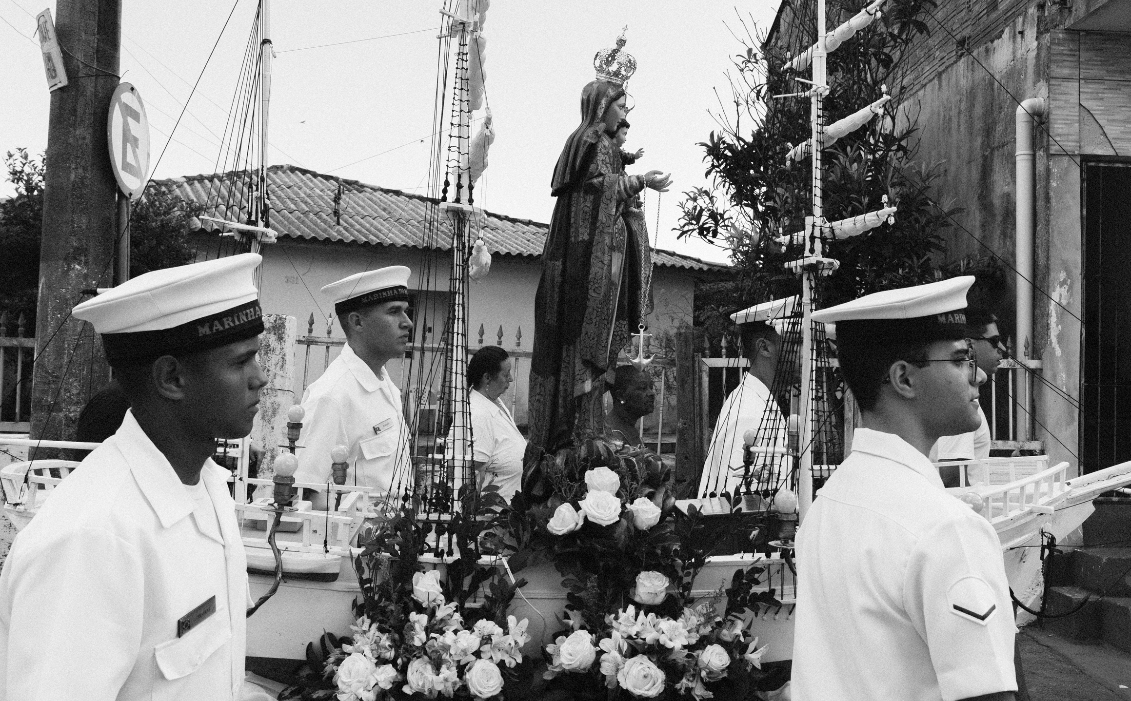 Imagem 6 da Galeria de Fotos. Audiodescrição: Fotografia em preto e branco, capturada em ambiente externo durante o dia, mostrando uma procissão religiosa com forte presença simbólica marítima. No centro da imagem está uma imagem de Nossa Senhora dos Navegantes com o Menino Jesus nos braços. A santa usa um manto ornamentado e uma grande coroa, e segura um escapulário com uma âncora pendurada, símbolo tradicional de proteção aos navegantes. A imagem sagrada está posicionada sobre uma base decorada que simula a estrutura de um navio, com mastros altos, cordames e velas enroladas. A base está ricamente ornada com flores claras, principalmente rosas brancas, que ocupam a parte inferior da embarcação devocional. Quatro homens com uniforme da Marinha — camisa branca de mangas curtas e quepe com a inscrição "MARINHA" — ladeiam a estrutura em posição de marcha, com semblantes sérios. Ao fundo, vê-se parte de uma casa com telhado de barro, um poste com placa de trânsito e o muro de outra construção. Uma mulher de roupas claras também caminha discretamente ao lado da imagem. A imagem transmite um forte sentido de tradição, devoção popular e sincretismo entre fé católica e cultura marítima, reforçando a reverência a Nossa Senhora dos Navegantes como padroeira dos pescadores e símbolo de esperança nas comunidades costeiras.