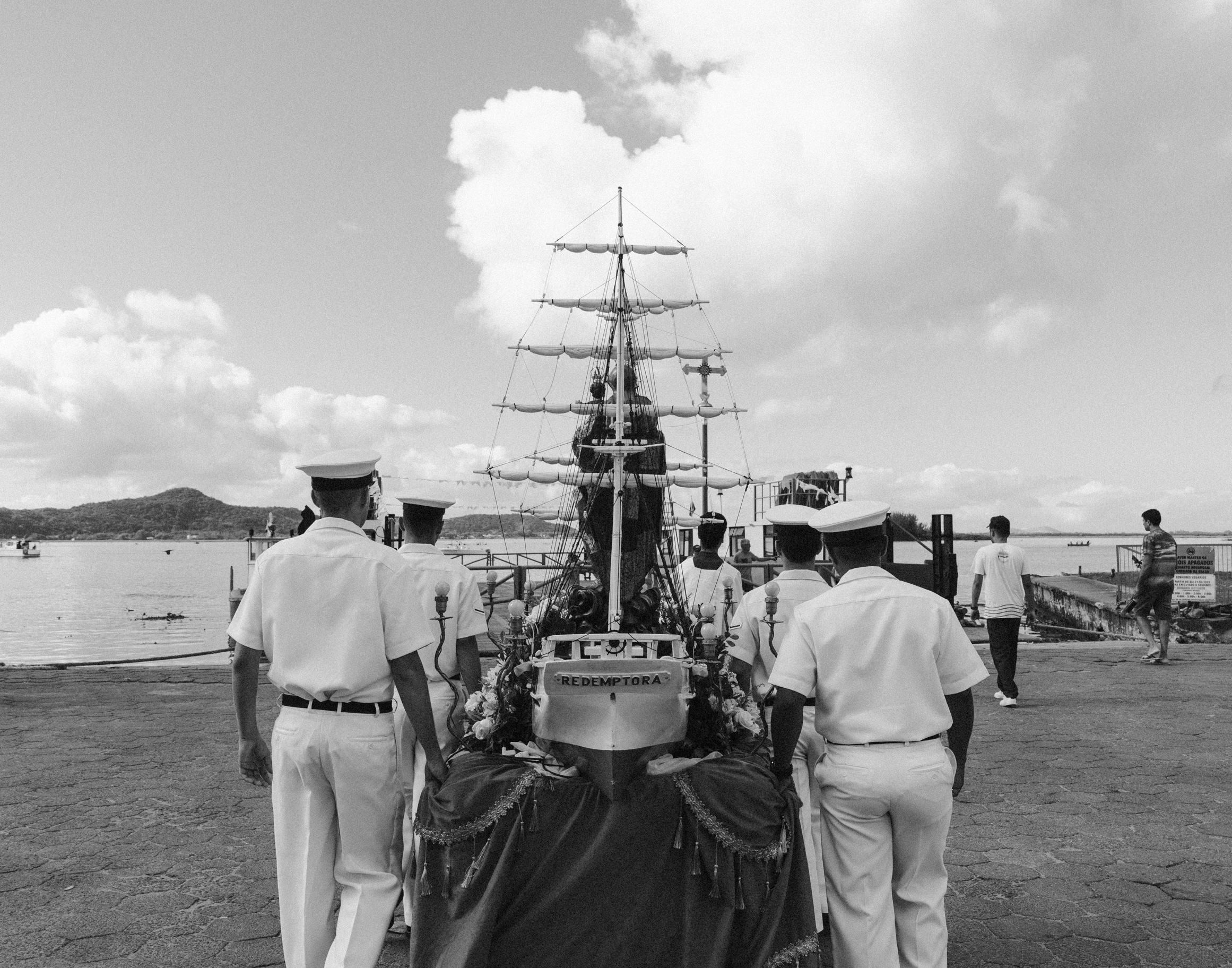 Imagem 7 da Galeria de Fotos. Audiodescrição: Fotografia em preto e branco, capturada em ambiente externo à luz do dia, mostrando uma procissão religiosa que se dirige a um cais à beira-mar. Em primeiro plano, vista de costas, está uma estrutura em formato de barco decorativo, com mastros altos, velas enroladas e cordames — simulando um navio antigo. No centro dessa estrutura está uma imagem de Nossa Senhora dos Navegantes, que usa um manto e uma coroa. A embarcação simbólica está adornada com flores e tecidos festivos, com uma faixa na parte de trás onde se lê a palavra "REDENTORA". A imagem está sendo transportada por homens uniformizados da Marinha do Brasil, todos usando trajes brancos e quepes, marchando solenemente em fila, escoltando o andor. Ao fundo, o mar calmo se estende até o horizonte, com alguns barcos pequenos flutuando e uma cadeia de morros em meio às nuvens. À direita da imagem, outras pessoas observam a cena ou caminham pelo cais. A atmosfera da imagem é de reverência, devoção e forte simbolismo cultural. A procissão segue em direção ao mar, para o embarque da imagem sagrada na festa religiosa tradicional de Nossa Senhora dos Navegantes.