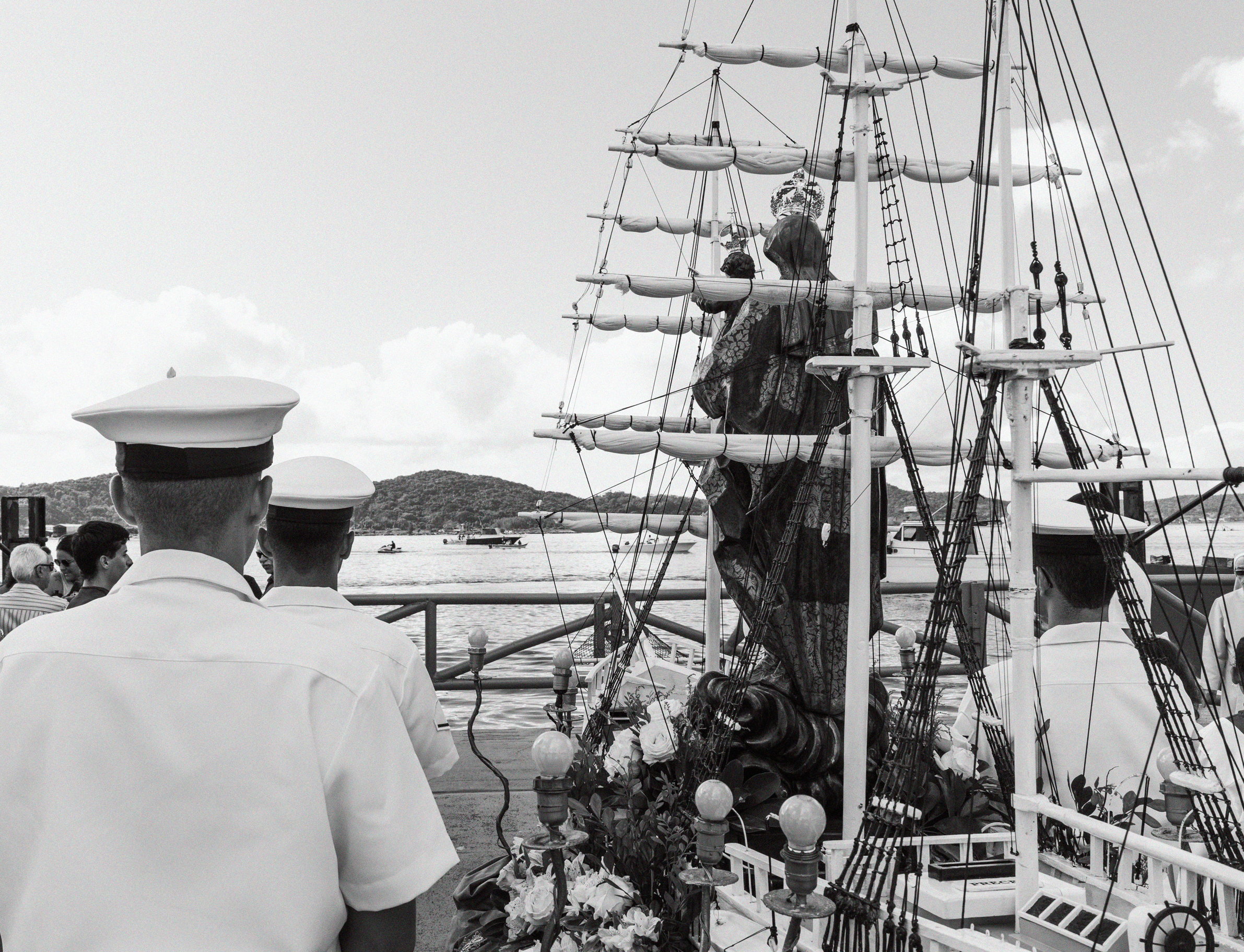Imagem 8 da Galeria de Fotos. Audiodescrição: Fotografia em preto e branco, capturada à luz do dia, retratando uma cerimônia religiosa à beira-mar. A cena mostra a imagem de Nossa Senhora dos Navegantes com o Menino Jesus nos braços, posicionada sobre uma base decorada que simula a estrutura de um navio antigo com mastros altos, cordames e velas enroladas. A escultura da santa está vestida com manto ornamentado e coroa, transmitindo solenidade e reverência. O Menino Jesus também usa uma pequena coroa. Em primeiro plano, à esquerda, dois marinheiros aparecem de costas, usando uniforme branco e quepes com aba escura. À direita, parcialmente visível, estão outros marinheiros e membros do cortejo. A base da escultura está ornada com flores e folhagens, bem como pequenos globos decorativos semelhantes a luminárias. Ao fundo, o mar calmo ocupa boa parte da imagem, com barcos e lanchas flutuando nas águas. Uma ilha ou colina coberta por vegetação preenche a linha do horizonte, sob um céu claro com nuvens suaves. A imagem transmite uma atmosfera de fé, devoção e tradição marítima, característica das celebrações dedicadas a Nossa Senhora dos Navegantes, padroeira dos pescadores. O contraste entre os elementos religiosos e o cenário natural costeiro acentua o simbolismo de proteção divina sobre os navegantes.