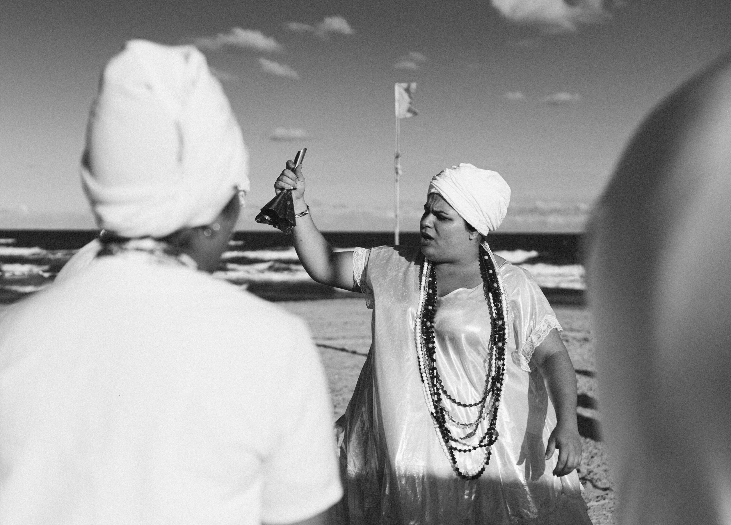 Galeria de Imagens. imagem 2: Imagem em preto e branco. Cena em uma praia, com o mar ao fundo e céu com poucas nuvens. Em primeiro plano, do lado esquerdo, há uma pessoa de costas, vestida de branco e usando um turbante. Em destaque, ao centro e à direita, está uma pessoa de frente, também com turbante branco e vestida com uma bata de tecido brilhante, possivelmente cetim. Ela segura um sino de metal com a mão direita erguida, em gesto expressivo, e parece estar falando. Usa colares longos e múltiplos, característicos de religiões de matriz africana. Ao fundo, vê-se uma bandeira em um mastro fincado na areia. A cena transmite um momento de ritual ou celebração religiosa à beira-mar.