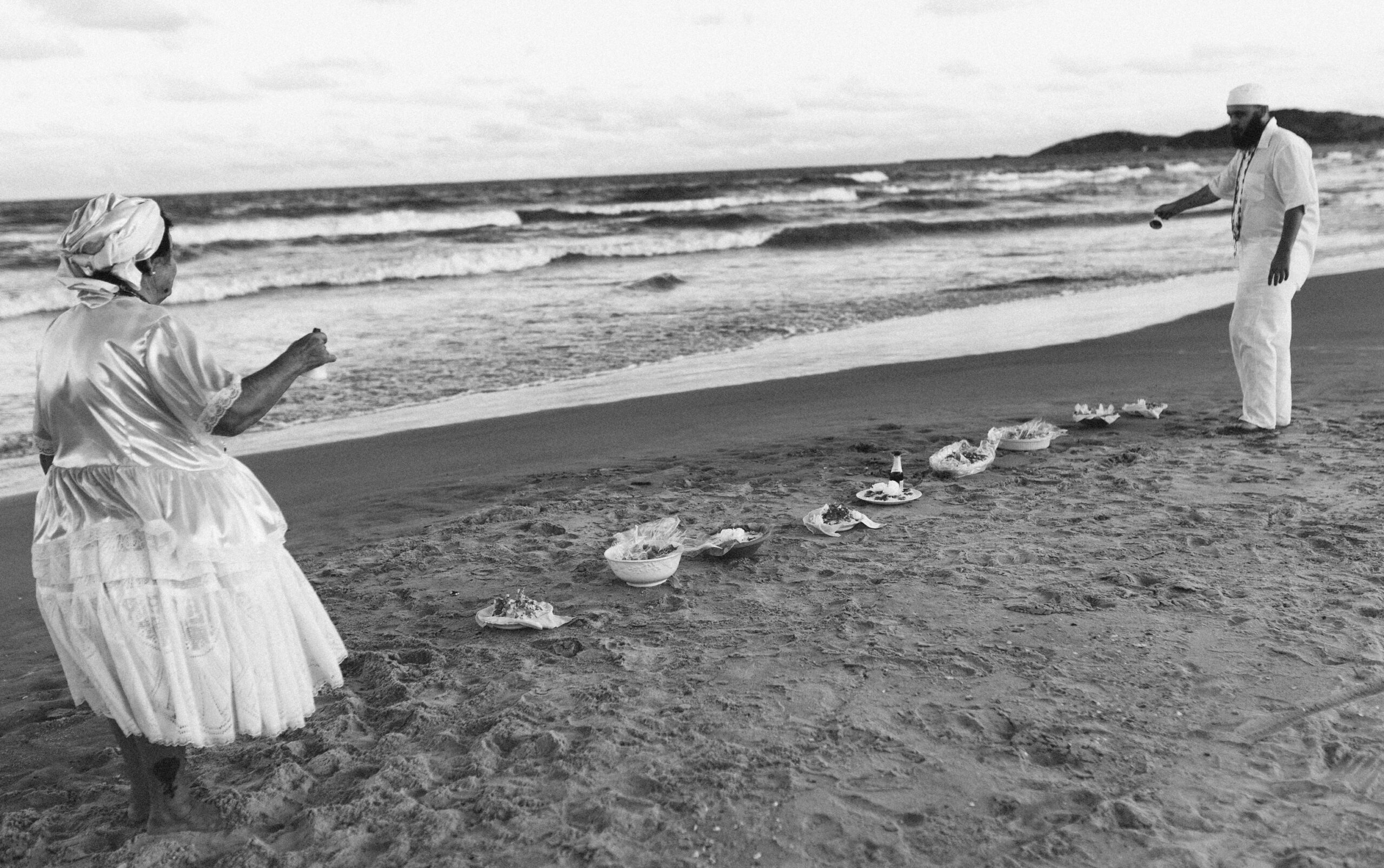 Galeria de Imagens. imagem 4: Imagem em preto e branco. Cena capturada à beira-mar, durante um ritual de umbanda. Em primeiro plano, duas pessoas estão de frente uma para a outra, de lados opostos da imagem. À esquerda, uma mulher de costas usa um vestido tradicional branco, de tecido brilhante e rendado, com saia volumosa e turbante na cabeça. À direita, um homem de frente segura um recipiente pequeno e parece despejar líquido na areia, em gesto ritualístico. Ele também está vestido de branco e usa colares longos no pescoço. Entre eles, dispostas em linha na areia, há várias oferendas: cestos e recipientes contendo alimentos, flores, folhas e objetos simbólicos. Ao fundo, o mar com ondas suaves e o céu parcialmente nublado. No horizonte, vê-se uma colina. A composição transmite espiritualidade, conexão com a natureza e reverência.