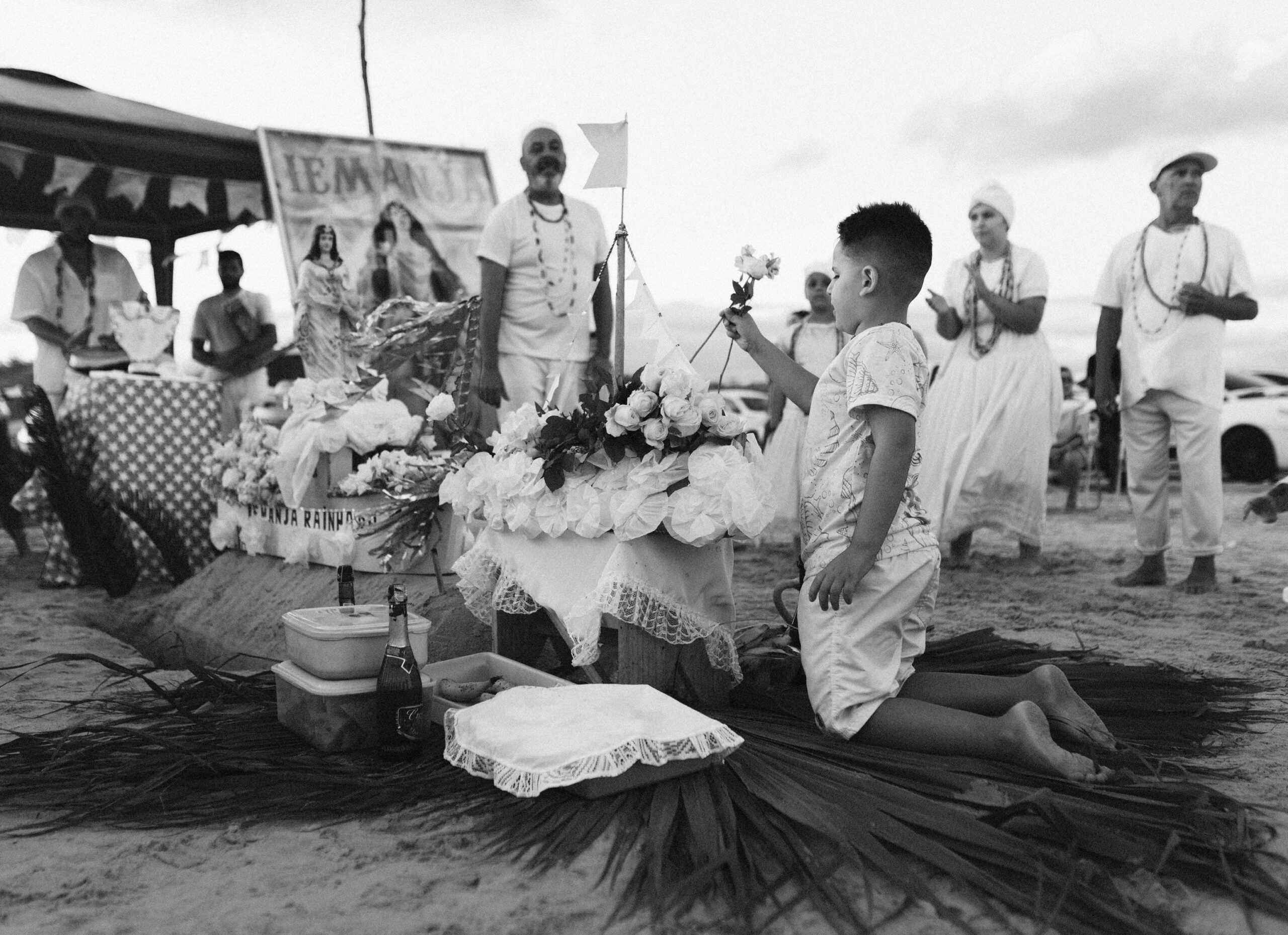 Galeria de Imagens. imagem 6: Imagem em preto e branco. A cena acontece sobre a areia da praia, com um grupo de pessoas reunidas ao redor de um altar decorado para Iemanjá, orixá das águas no candomblé e na umbanda. Em destaque, no canto direito da imagem, um menino de joelhos está voltado para o altar, segurando uma flor branca estendida em direção à oferenda. Ele veste uma camiseta clara com desenhos e bermuda branca. O altar é ricamente decorado com flores brancas, papel rendado, tecidos e uma pequena embarcação com uma vela branca hasteada. Há também uma estátua de Iemanjá e um banner ao fundo com a palavra “IEMANJÁ” escrita em letras grandes, além de imagens da divindade. Sobre o altar e no chão ao redor, há garrafas de bebidas e folhas de palmeira dispostas como uma base cerimonial. Ao fundo, outras pessoas observam e participam da cerimônia, vestidas em trajes brancos e colares de contas, típicos de religiões de matriz africana.