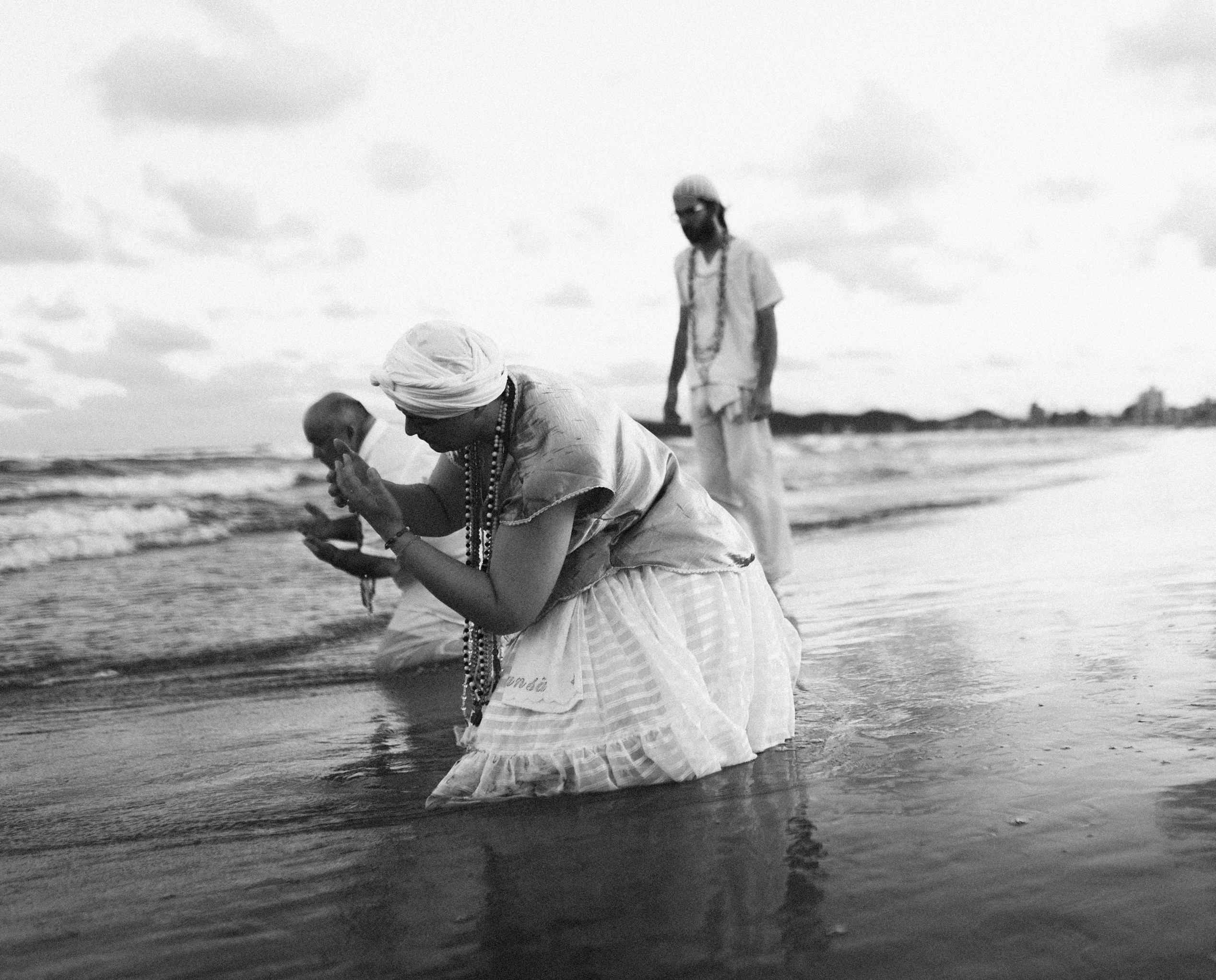 Galeria de Imagens. imagem 8: Imagem em preto e branco. A fotografia mostra três pessoas à beira-mar participando de um ritual religioso, ligado à umbanda. Em destaque, no primeiro plano, uma mulher está ajoelhada na água com o corpo inclinado para frente e as mãos em posição de oração ou oferenda. Ela veste uma saia rodada branca com detalhes em renda, blusa clara e turbante branco. Vários colares coloridos pendem do seu pescoço, representando elementos de devoção. À esquerda, outro participante, também ajoelhado, realiza um gesto similar com as mãos em concha, próximo à superfície da água, possivelmente em um momento de purificação. Ao fundo, uma terceira pessoa está em pé, observando. Todos estão vestidos com roupas brancas, típicas dos rituais de religiões afro-brasileiras. O mar calmo reflete parte das figuras, e ao longe vê-se a linha do horizonte, com pequenas elevações e edifícios à direita. O céu está parcialmente nublado, criando uma atmosfera serena e sagrada. A imagem transmite espiritualidade, entrega e conexão com a natureza, em especial com o mar — elemento essencial nos cultos a Iemanjá, orixá das águas.