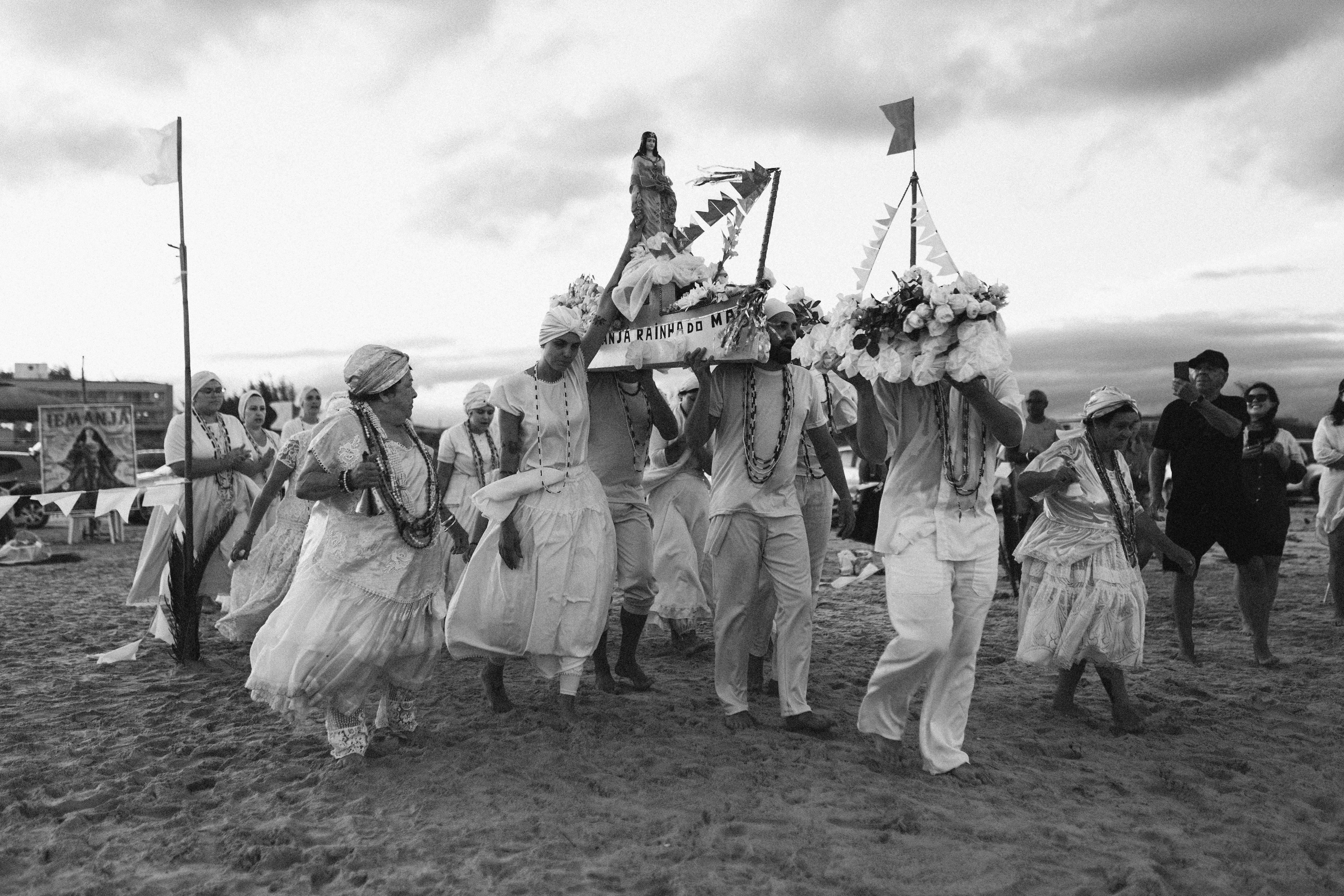 Audiodescrição da Imagem: Imagem em preto e branco. A fotografia registra um grupo de pessoas participando de um cortejo religioso em uma praia, provavelmente em uma cerimônia em homenagem a Iemanjá. O grupo caminha descalço sobre a areia. Ao centro da imagem, dois homens carregam sobre os ombros uma espécie de andor decorado com flores, fitas e bandeirinhas. No topo, há uma imagem da divindade Iemanjá e uma faixa com os dizeres "RAINHA DO MAR". O cortejo é composto majoritariamente por mulheres vestidas com roupas brancas tradicionais, saias rodadas e turbantes,adornadas com colares típicos das religiões de matriz africana. Uma senhora mais velha caminha à frente, com expressão solene, segurando sua saia enquanto se movimenta. No fundo, pessoas observam e registram o momento com celulares. À esquerda, vê-se uma tenda decorada com bandeirinhas e um cartaz com a imagem de Iemanjá e seu nome escrito em letras grandes. O céu está parcialmente nublado, com nuvens densas e luz suave, sugerindo o entardecer.