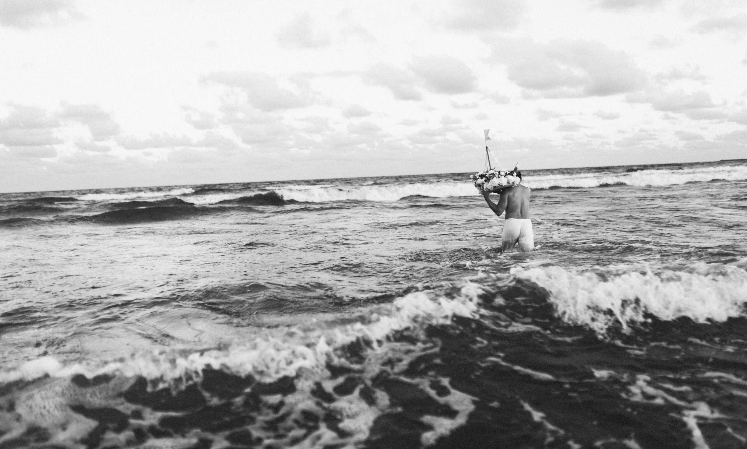 Galeria de Imagens. imagem 10: Imagem em preto e branco. Fotografia capturada em plano geral e em ângulo ligeiramente baixo, mostrando uma pessoa sozinha entrando no mar. A cena é registrada de costas. A pessoa está imersa até a metade das coxas na água revolta e caminha em direção ao horizonte, com ondas quebrando ao seu redor. Ela veste uma calça clara e carrega sobre o ombro esquerdo uma pequena embarcação decorada com flores e bandeirolas onde tem uma pequena escultura de Iemanjá. O céu está parcialmente nublado, com nuvens espalhadas e o mar se estende até a linha do horizonte. A atmosfera da imagem é contemplativa e simbólica, sugerindo um momento ritualístico ou de conexão espiritual com a natureza.