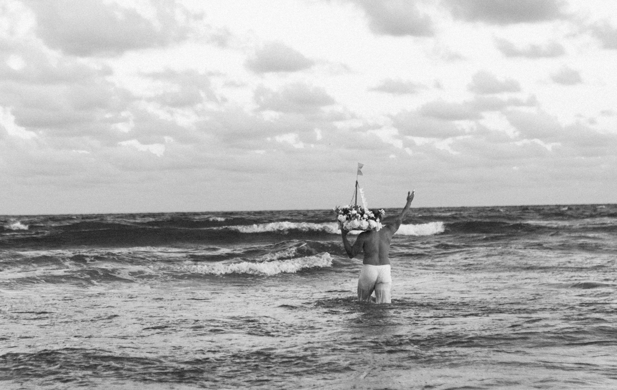 Galeria de Imagens. imagem 11: Imagem em preto e branco. Fotografia capturada em plano geral onde um homem de costas caminha mar adentro, com a água chegando até pouco acima dos joelhos. Ele veste uma calça clara e está com o torso nu. Em seu ombro esquerdo, uma pequena embarcação decorada com flores e bandeirolas onde tem uma pequena escultura de Iemanjá. O objeto possui estrutura triangular e está ornado com uma faixa de tecido que lembra uma vela, além de uma pequena bandeira no topo. O homem ergue o braço direito em direção ao céu, com a mão aberta e os dedos apontando para cima, gesto que pode ser interpretado como uma saudação, oferenda ou invocação simbólica. O mar apresenta ondas agitadas, e o céu parcialmente nublado preenche a parte superior da imagem, reforçando a atmosfera contemplativa e ritualística da cena.