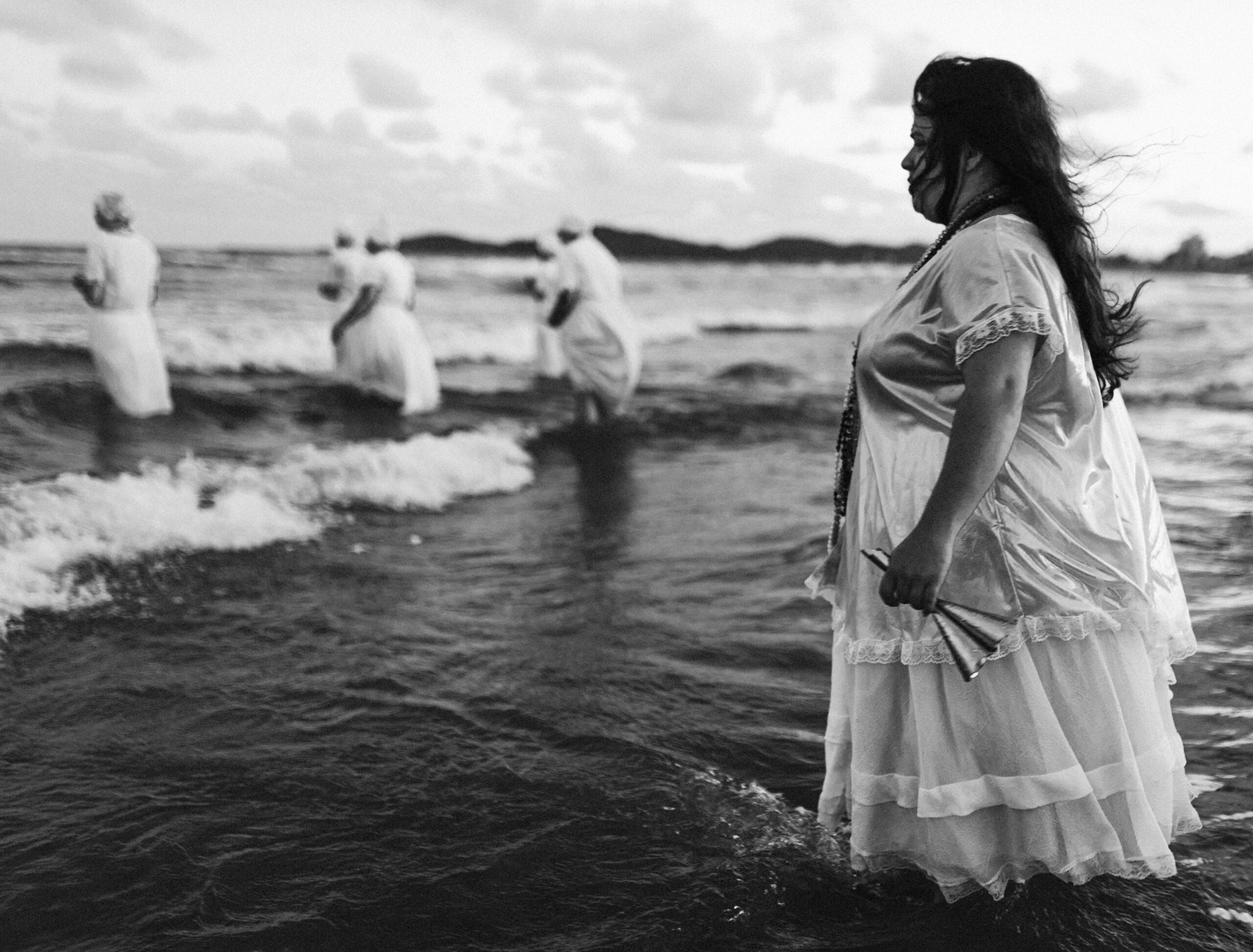 Galeria de Imagens. imagem 13: Fotografia em preto e branco, tirada em plano geral com foco na lateral direita, retratando um grupo de mulheres dentro do mar raso, em uma cena com forte conotação ritualística. Em primeiro plano, à direita da imagem, uma mulher de perfil olha para o horizonte. Ela veste um vestido branco longo e rodado, com acabamento em renda, e segura uma sineta em uma das mãos. Seus cabelos escuros estão soltos e esvoaçantes ao vento, e ela usa colares longos no pescoço. Ao fundo, outras cinco mulheres, também de branco e com saias volumosas, caminham mar adentro. Algumas usam turbantes na cabeça. A maioria está de costas ou levemente de lado, participando de uma oferenda no mar. O céu está parcialmente nublado, com nuvens espalhadas, e o horizonte é marcado por uma faixa de terra ao fundo. As ondas do mar são pequenas, mas contínuas, e tocam suavemente as vestes das mulheres. A cena transmite um sentimento de espiritualidade, respeito e conexão com elementos da natureza e da fé.