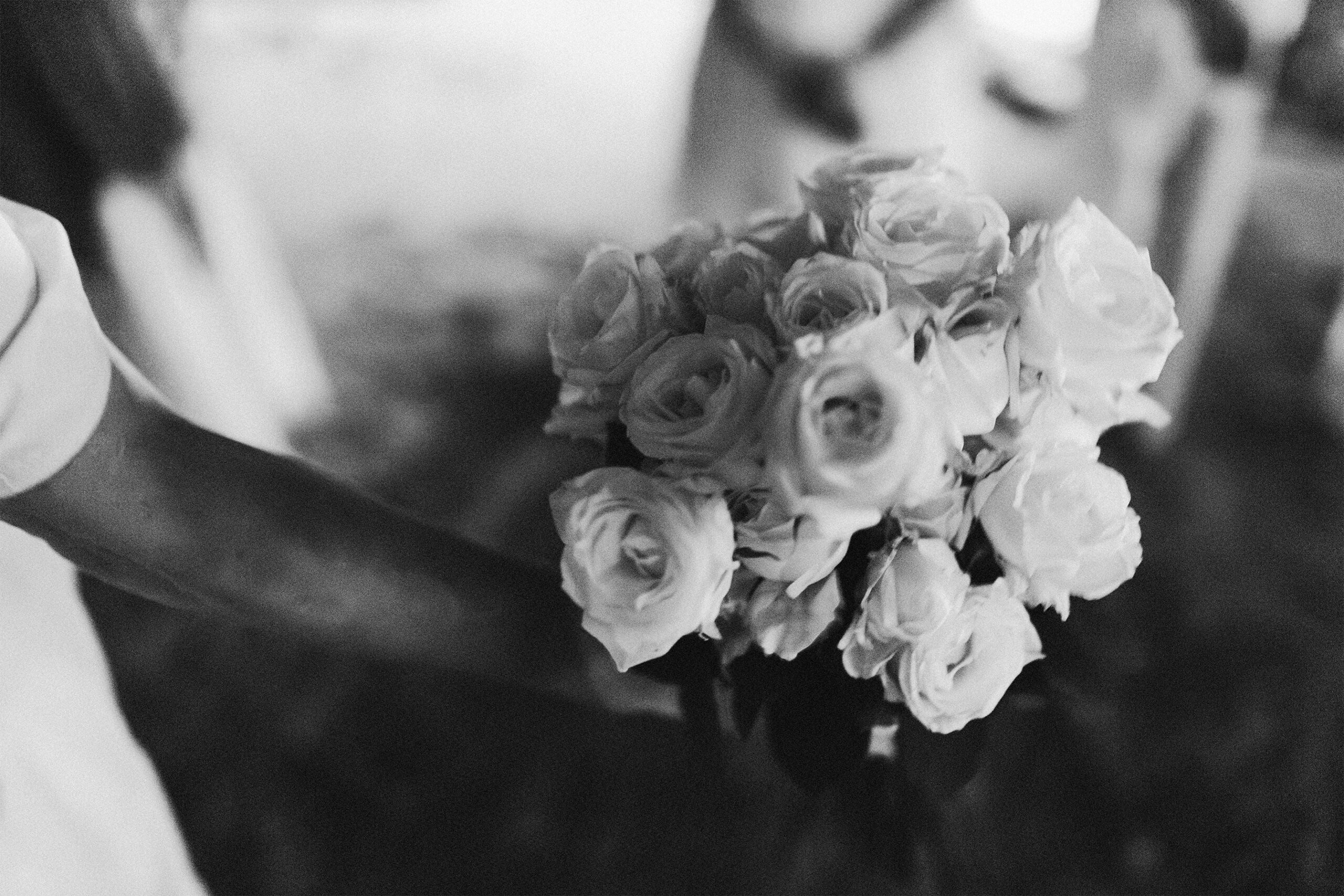 Galeria de Imagens. imagem 29: Fotografia em preto e branco, capturada em plano detalhe e em foco seletivo no tema. A imagem mostra um buquê de rosas brancas sendo segurado por uma pessoa à esquerda da cena, cujo braço e parte da roupa são visíveis. O buquê é composto por diversas rosas em diferentes estágios de abertura, formando uma composição volumosa e delicada. A mão que o segura está parcialmente fora de foco, assim como o fundo da imagem, onde se percebem silhuetas humanas e formas indefinidas, sugerindo um ambiente com outras pessoas. A textura suave das pétalas contrasta com a leve granulação da imagem. A cena transmite sensibilidade, reverência e simbolismo, com forte conotação afetiva ou espiritual.