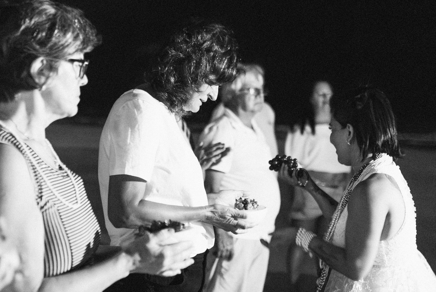 Galeria de Imagens. imagem 15: Fotografia em preto e branco, tirada em plano médio, capturando um grupo de pessoas participando de um ritual noturno à beira-mar. A cena é iluminada por uma fonte de luz artificial intensa, que destaca os rostos e os objetos nas mãos dos participantes. Em primeiro plano, à direita, uma mulher com vestido rendado e diversos colares no pescoço — características comuns a trajes religiosos e adereços das religiões afro-brasileiras — segura uvas e interage com as demais pessoas. À esquerda, outras mulheres, algumas segurando pequenos recipientes brancos com o que parecem ser flores olham atentamente para a cerimônia. Os trajes variam entre camisetas brancas e roupas listradas, com predominância do branco. Ao fundo, mais participantes observam a cena, todos em postura respeitosa e atenta. O fundo é escuro, com pouca definição, sugerindo que o ritual ocorre durante a noite na praia. A imagem transmite uma atmosfera solene, de espiritualidade coletiva.