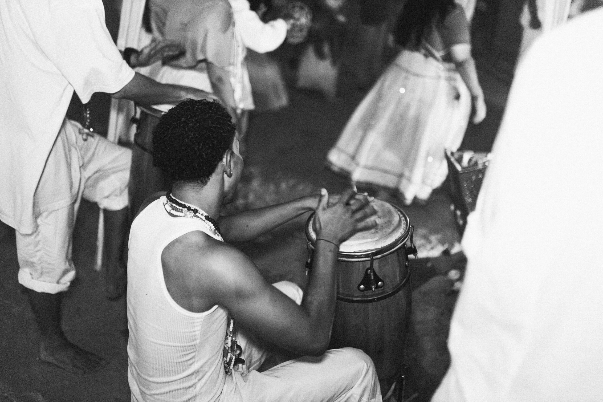 Imagem 7. Audiodescrição: Imagem em preto e branco. Fotografia capturada em plano médio e em perspectiva lateral. Em primeiro plano, ao centro da imagem, está um homem negro sentado, visto de costas e ligeiramente de perfil, tocando um atabaque — instrumento de percussão típico das religiões de matriz africana. Ele veste uma regata branca e calça clara, e tem colares religiosos pendendo do pescoço. Seus braços estão erguidos, em movimento, no momento em que as mãos tocam a pele do tambor, transmitindo dinamismo à cena. Ao redor, outros participantes da cerimônia estão parcialmente visíveis. À esquerda, um homem em pé toca outro instrumento de percussão; à direita, uma mulher com saia longa e rodada, bordada e clara, caminha de costas. Ao fundo, outras pessoas completam a composição, algumas em trajes brancos, outras com elementos rituais. O chão é de areia da praia e o evento ocorre à noite. A imagem expressa movimento, ritmo e espiritualidade, evocando uma cerimônia coletiva onde música e devoção se entrelaçam num ambiente de celebração ancestral.