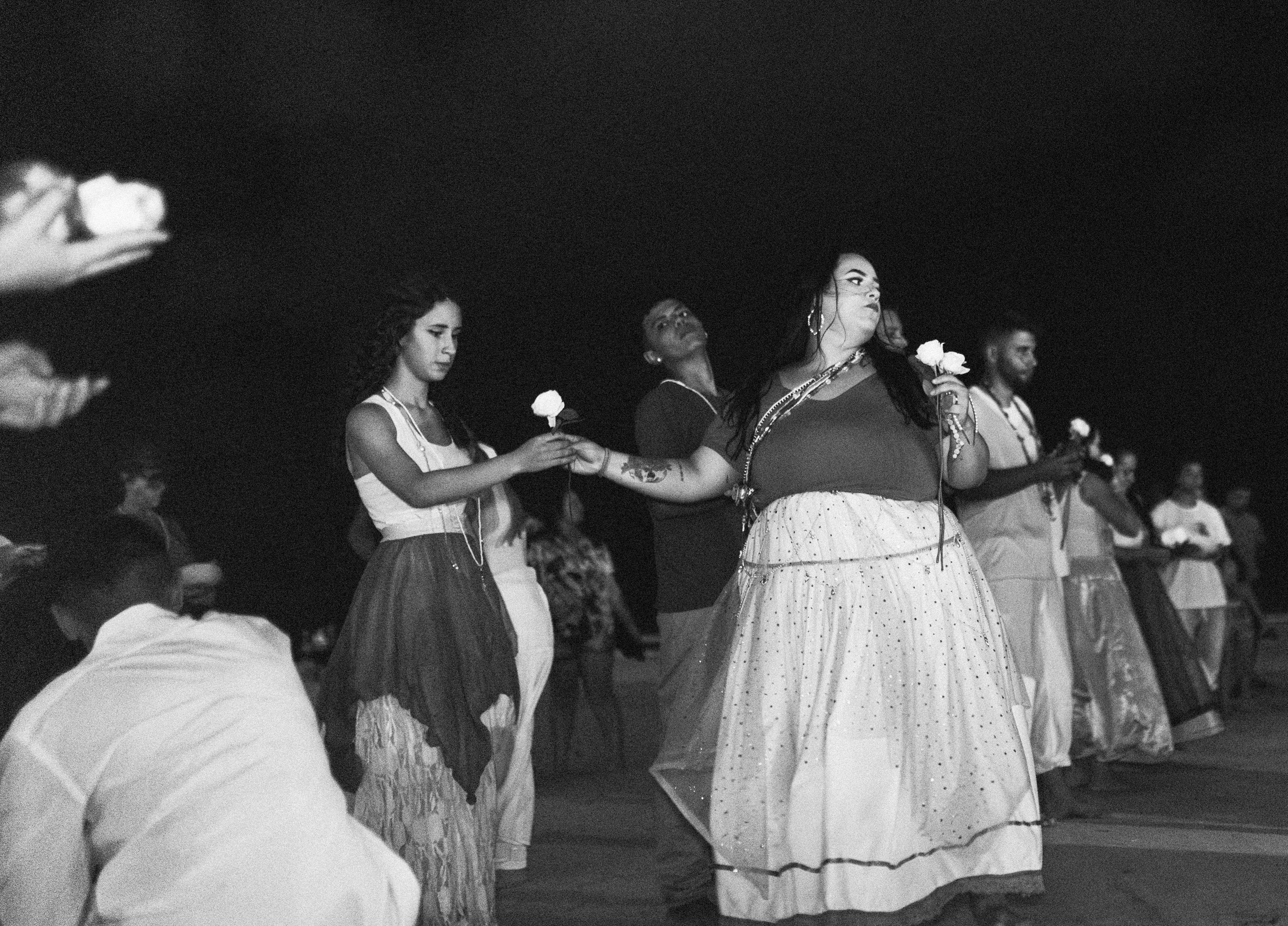 Galeria de Imagens. imagem 17: Imagem em preto e branco. Fotografia capturada em plano geral e em ambiente noturno, retratando um grupo de pessoas participando de um ritual coletivo ao ar livre em um contexto religioso de matriz africana. A cena se passa com o céu escuro ao fundo. No centro da imagem, duas mulheres se destacam: uma jovem de cabelo encaracolado, vestindo blusa clara, saia longa fluida em camadas e colares, estende uma rosa branca a uma mulher maior, que usa blusa escura e uma saia longa bordada com pequenos brilhos. A mulher que recebe a rosa segura outras flores na mão esquerda e está adornada com colares e pulseiras, com o olhar voltado para o lado direito da imagem, demonstrando concentração ou solenidade. Ao fundo, outros participantes, homens e mulheres, formam uma fileira. Muitos seguram rosas brancas, elemento recorrente em rituais de oferenda. As vestes são predominantemente claras, com tecidos leves e adornos religiosos. Algumas pessoas usam colares típicos das tradições afro-brasileiras. À esquerda, mãos de outros participantes também aparecem segurando flores, reforçando a ação coletiva. A cena transmite um forte sentido de união, espiritualidade e respeito, num momento de entrega simbólica e conexão com o sagrado.