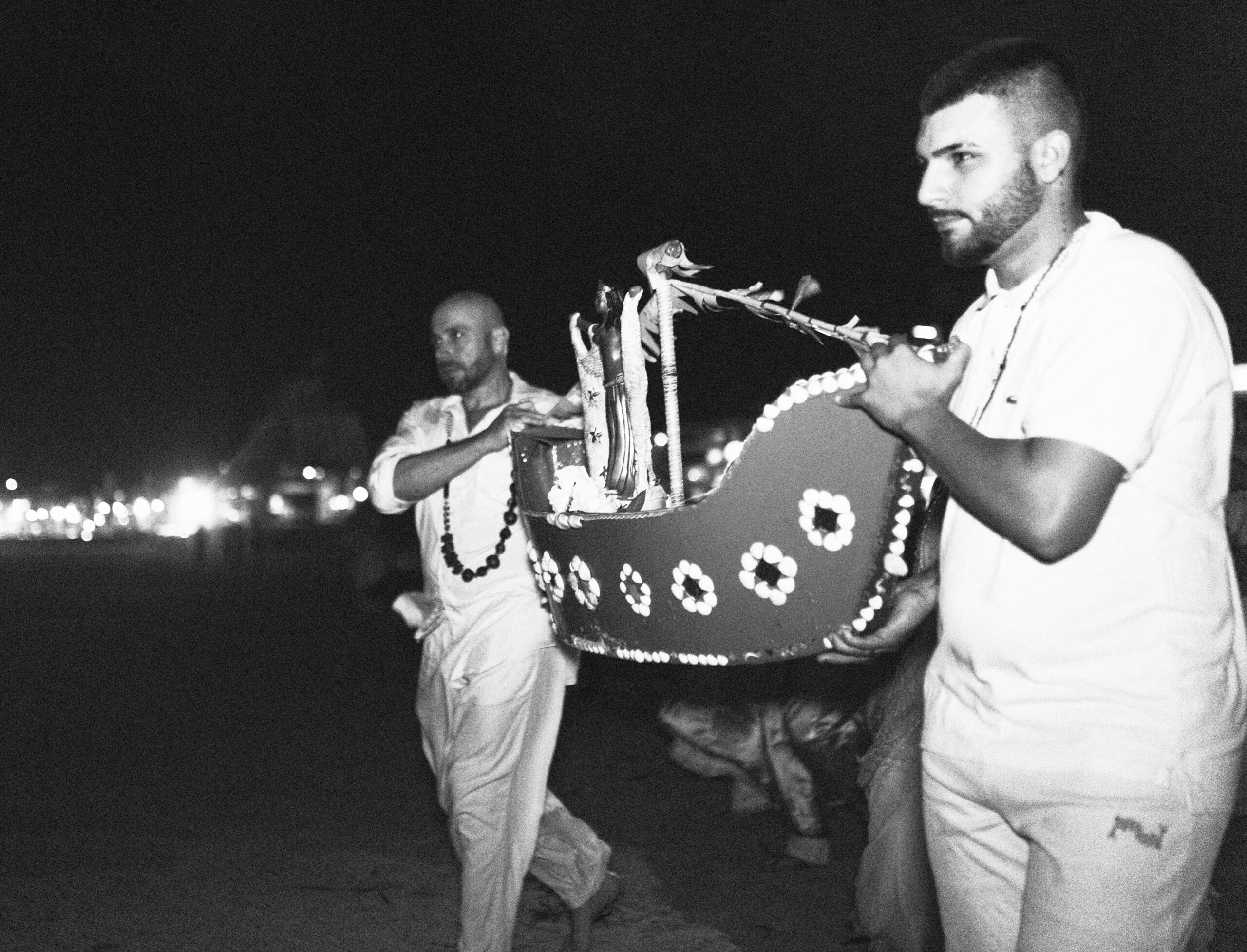 Galeria de Imagens. imagem 18: Fotografia em preto e branco, capturada em plano médio e em ambiente noturno, mostrando dois homens carregando uma peça ritualística em formato de barco estilizado, decorada com flores e enfeites. A embarcação parece feita de material leve e tem formato curvo, com detalhes circulares ao longo da borda, que se destacam pela ornamentação em alto contraste. Há flores dentro do barco. Os homens caminham lado a lado. O homem à direita, em primeiro plano, é branco, tem cabelo curto e barba aparada, usa camiseta clara e colares rituais. Ele segura a parte dianteira da embarcação com ambas as mãos. O homem à esquerda, ligeiramente atrás, é negro, possui cabelo raspado, usa camisa de manga comprida arregaçada e calça branca. Também carrega colares religiosos e segura a extremidade traseira do barco. Ao fundo, à esquerda, percebe-se um ponto de luz intensa e várias luzes pequenas, sugerindo a presença de uma cidade ou vila litorânea. O chão é de areia, e o céu completamente escuro.