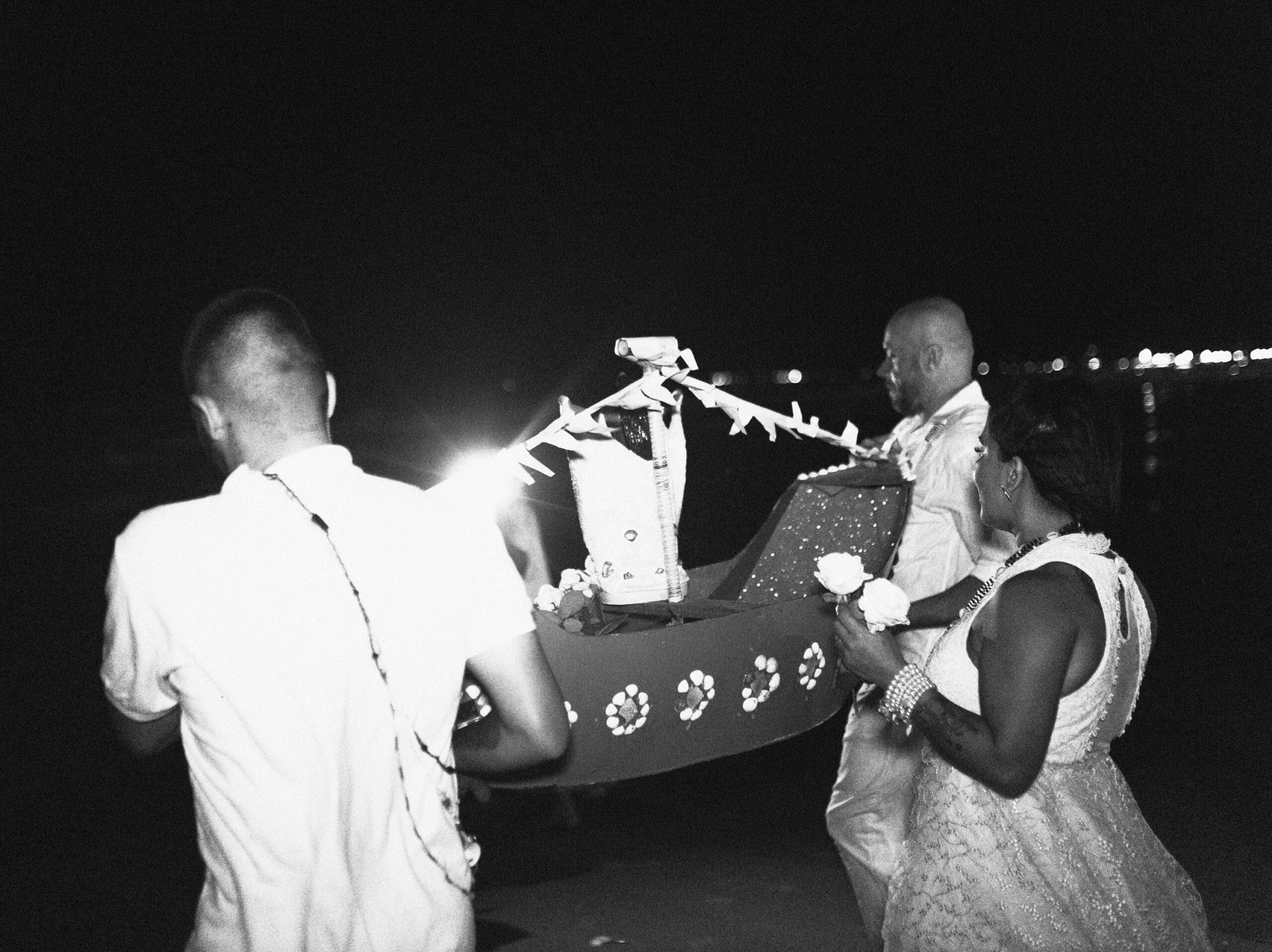 Galeria de Imagens. imagem 19: Fotografia em preto e branco, capturada em plano médio e em ambiente noturno, retratando um momento ritualístico com forte carga simbólica. No centro da imagem, três pessoas carregam uma embarcação artesanal decorada, representando um barco cerimonial. O objeto é adornado com conchas e flores dispostas em padrões circulares e contém no seu interior uma imagem representando Iemanjá. À esquerda, um homem de costas, com cabelo raspado, veste camisa clara e um colar religioso que cai pelas costas. Ele segura a parte traseira da embarcação. Do lado oposto, outro homem de costas e trajando branco, segura a parte frontal do barco com ambas as mãos. À direita da imagem, uma mulher com vestido rendado claro, colares e pulseiras brancas segura duas rosas brancas, caminhando ao lado do barco. Ela observa a embarcação com expressão solene. Ao fundo, o mar e a linha de costa aparecem escurecidos, iluminados apenas por pontos de luz ao longe e por um feixe forte vindo da direção oposta à câmera, que destaca os participantes e o barco.