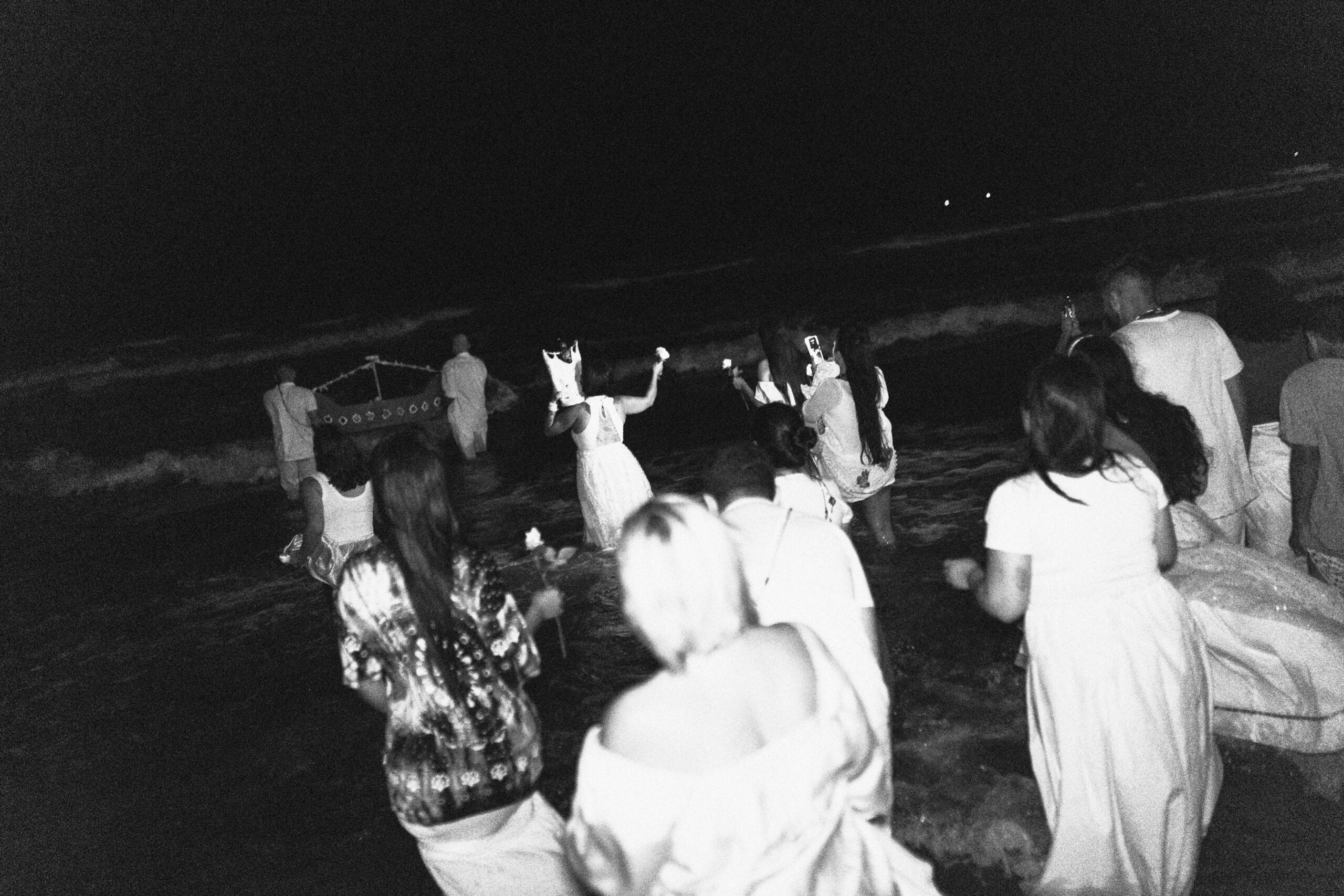 Galeria de Imagens. imagem 20: Fotografia em preto e branco, capturada em plano geral e ambiente noturno, mostrando um grupo de pessoas dentro do mar, participando de um ritual coletivo de oferenda a Iemanjá. A cena é registrada com as pessoas de costas caminhando em direção ao mar. O mar está agitado, com ondas visíveis, e o céu escuro ao fundo cria um contraste com as vestes claras dos participantes. Em destaque, no centro da imagem, duas pessoas — uma à esquerda e outra à direita — carregam uma embarcação artesanal decorada, semelhante a um barco estilizado. Outros participantes os acompanham, alguns segurando flores, especialmente rosas brancas. Muitas pessoas estão parcialmente submersas até os joelhos ou coxas, em trajes predominantemente brancos ou claros, com algumas exceções que vestem roupas estampadas. À esquerda, uma mulher de vestido rendado e cabelo solto ergue um objeto acima da cabeça — possivelmente uma pequena escultura ritualística. Outras pessoas estão fotografando ou filmando o momento com celulares, o que demonstra a importância simbólica e emocional da ocasião. A atmosfera da imagem é carregada de forte senso de devoção e conexão com as águas do mar.