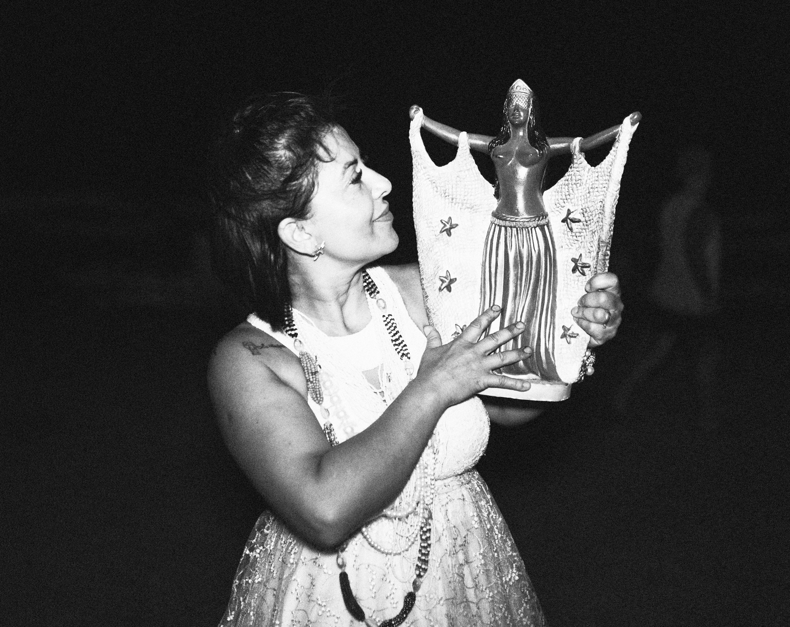 Galeria de Imagens. imagem 21: Fotografia em preto e branco, capturada em plano médio e ambiente noturno. No centro da imagem, uma mulher está em pé, de frente, segurando com ambas as mãos uma escultura que representa a figura feminina de Iemanjá. A escultura é negra, tem braços abertos, véus esvoaçantes com estrelas do mar em relevo e uma longa saia, com expressão serena. A mulher que segura a imagem tem pele clara e cabelos escuros presos para trás, sorri levemente enquanto olha com devoção para a imagem que segura próxima ao rosto. Ela usa um vestido branco rendado e adornos típicos de rituais religiosos afro-brasileiros: colares longos, pulseiras e um tecido amarrado à cintura. Ao fundo, o ambiente é escuro, indicando que a cena ocorre à noite. A imagem transmite um momento de fé, reverência e conexão espiritual, com destaque para a relação entre a mulher e a representação simbólica que ela carrega com carinho.