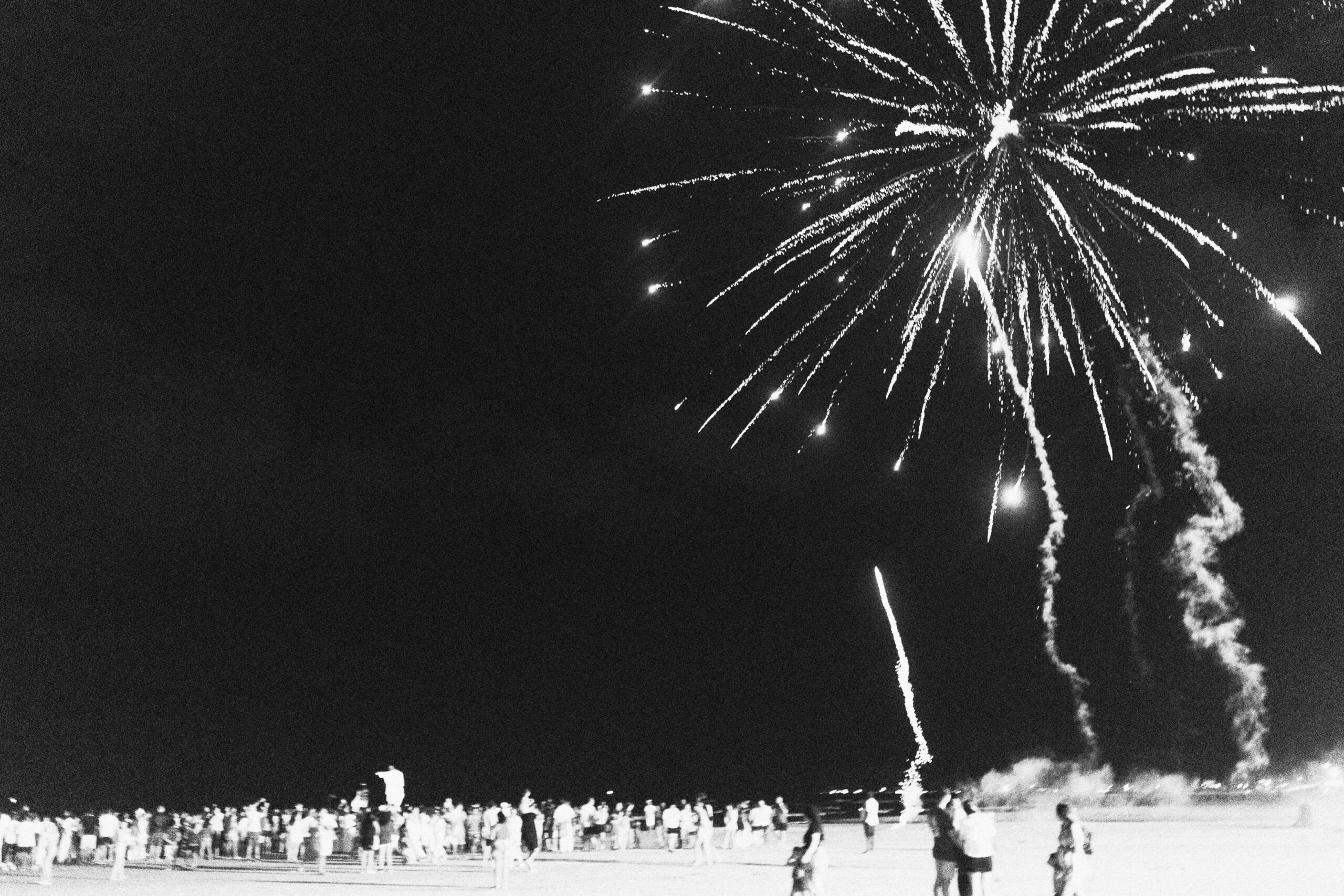 Galeria de Imagens. imagem 22: Fotografia em preto e branco, capturada em plano aberto e em ambiente noturno, retratando uma multidão reunida na praia sob um céu escuro, iluminado por fogos de artifício. No canto superior direito, os fogos explodem em feixes luminosos que se espalham em várias direções, formando um grande espetáculo visual. Trilhas de fumaça onduladas descem do céu, visíveis contra o fundo escuro. Na faixa inferior da imagem, uma grande quantidade de pessoas está distribuída ao longo da areia, observando os fogos. Algumas caminham, outras estão paradas, e várias figuras aparecem apenas como silhuetas ou vultos devido à iluminação de fundo. A claridade intensa próxima ao solo, provavelmente proveniente dos próprios fogos ou de refletores, destaca parcialmente a multidão. Ao fundo, no canto esquerdo, percebe-se uma leve linha de luzes da cidade à distância, indicando que a celebração ocorre em um local litorâneo habitado.