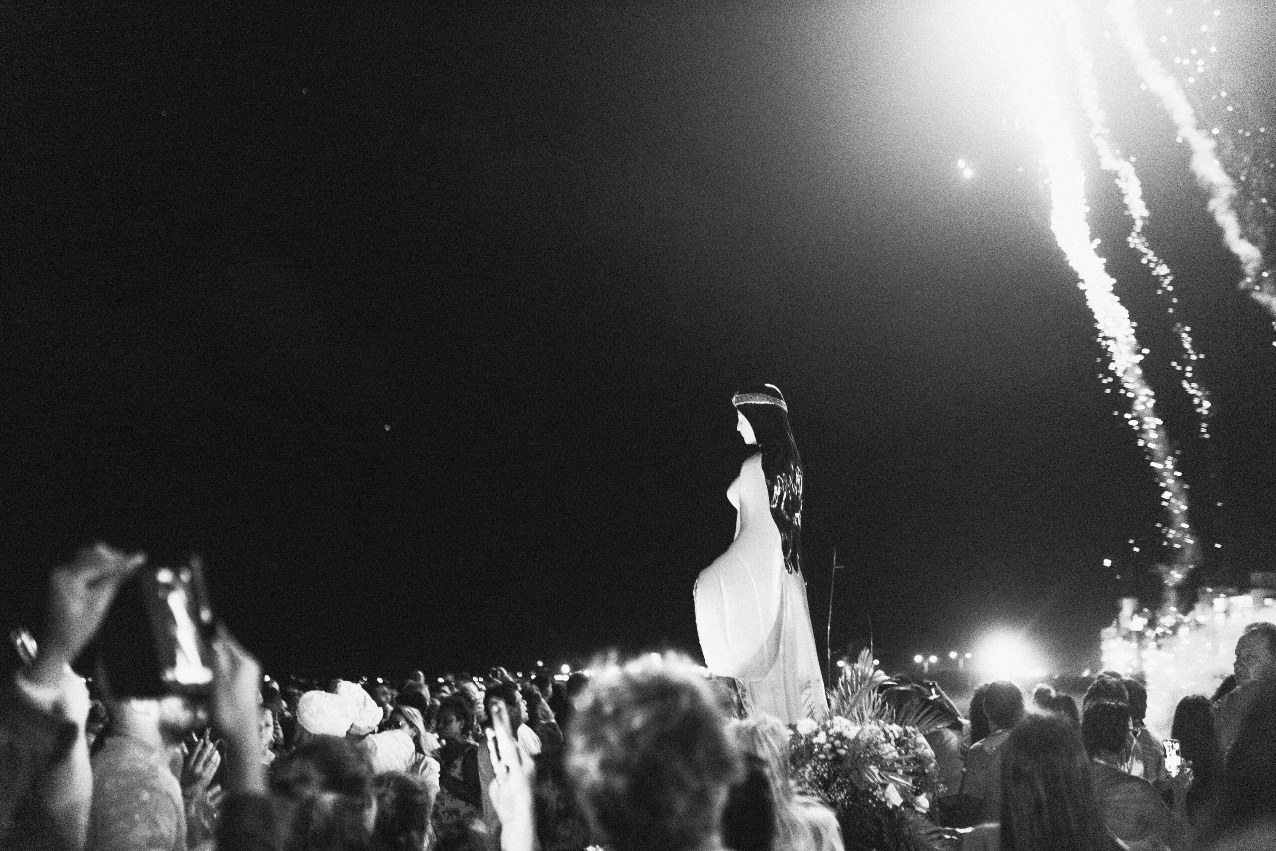 Imagem 5. Audiodescrição: Audiodescrição da Imagem: Fotografia em preto e branco, capturada em plano aberto durante a noite, com foco em uma multidão reunida ao ar livre. No centro da composição, erguida sobre uma base ornamentada com flores e folhagens, está uma grande estátua representando Iemanjá. A figura feminina está voltada para a direita da imagem, com os longos cabelos escuros caindo pelas costas e um vestido esvoaçante que acentua seu aspecto sereno e protetor. À frente da estátua, várias pessoas se aglomeram, algumas com as mãos erguidas, outras segurando celulares para registrar o momento. Muitas usam roupas claras e trajes tradicionais, compondo um ambiente devocional e festivo. No céu escuro ao fundo, fogos de artifício riscam a noite, com rastros de luz e explosões brilhantes no canto superior direito da imagem, reforçando o caráter celebrativo da ocasião. A atmosfera geral é de reverência, emoção coletiva e festa. A estátua de Iemanjá, imponente e iluminada, domina a cena como símbolo central de fé e proteção.