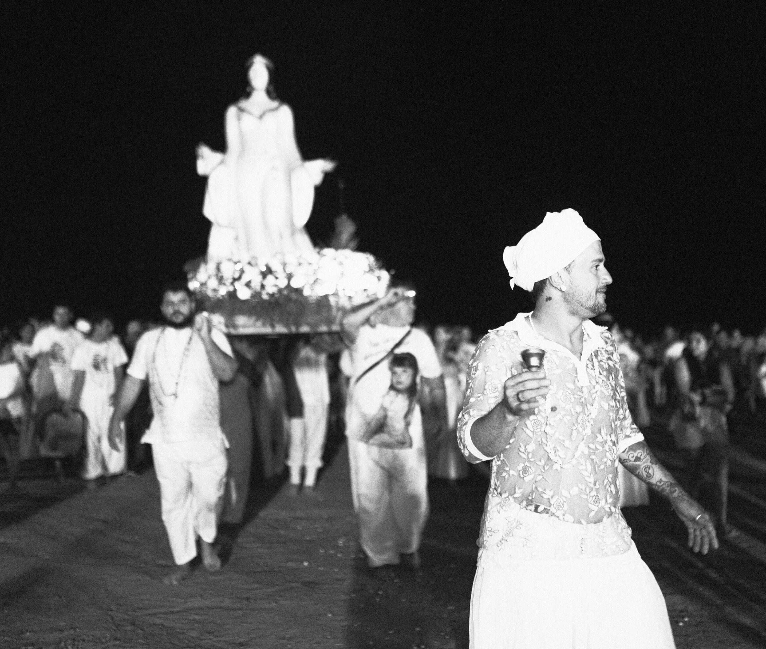 Galeria de Imagens. imagem 24: Fotografia em preto e branco, registrada em plano geral e ambiente noturno, mostrando uma procissão religiosa na areia de uma praia. No centro da imagem, uma grande imagem de Iemanjá — representada como uma figura feminina de braços abertos, cabelos longos e vestes esvoaçantes — é transportada sobre uma plataforma ornamentada com flores e luzes. A escultura está erguida acima da multidão, carregada nos ombros por ao menos quatro homens, dois visíveis em destaque na imagem. À frente da procissão, no lado direito da composição, um homem branco caminha segurando um pequeno sino ritualístico. Ele veste uma camisa de renda translúcida, calça branca e um turbante amarrado na cabeça. Seu olhar está voltado para a lateral direita da imagem. Ao fundo, uma multidão de pessoas acompanha o cortejo, muitas usando roupas claras ou brancas, algumas segurando flores ou objetos simbólicos. O céu está completamente escuro, destacando a cena iluminada por luz artificial.