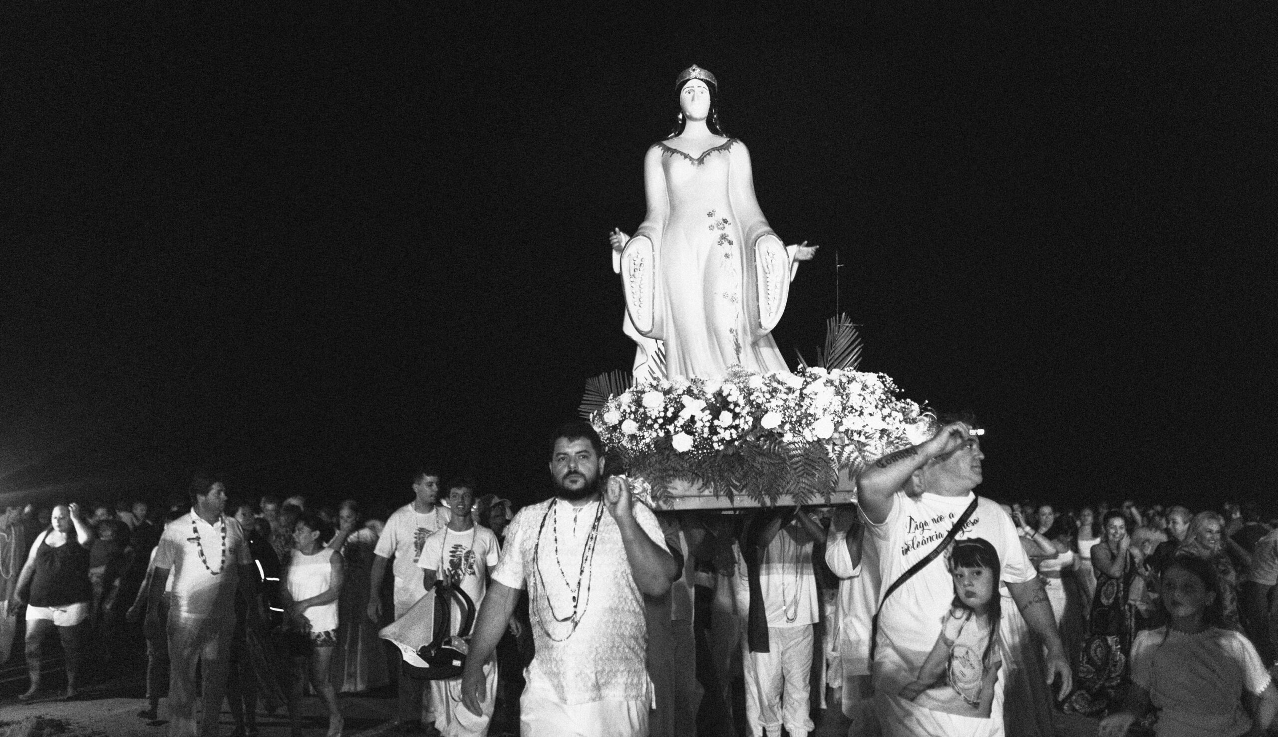 Galeria de Imagens. imagem 25: Fotografia em preto e branco, registrada em plano geral e ambiente noturno, retratando uma procissão em homenagem a Iemanjá. Ao centro da imagem, uma grande estátua da divindade é carregada por um grupo de homens. A escultura representa uma mulher de longos cabelos escuros, vestindo túnica esvoaçante e coroa na cabeça, com os braços estendidos para frente em gesto acolhedor. A base que sustenta a imagem está ricamente ornamentada com flores brancas e folhagens. Dois homens estão em primeiro plano, nas laterais da estrutura de madeira que sustenta a imagem. À esquerda, um homem branco, de barba, usando roupa branca rendada e colares religiosos. À direita, outro homem, também branco, segura a base com uma das mãos. Atrás deles, muitos outros carregadores e participantes acompanham a procissão. A multidão ao fundo é composta por pessoas de diferentes idades, todas acompanhando o cortejo com expressões de devoção e respeito. A maioria veste roupas brancas. O céu completamente escuro destaca a imagem iluminada da orixá, que se ergue sobre a multidão como figura central da celebração.