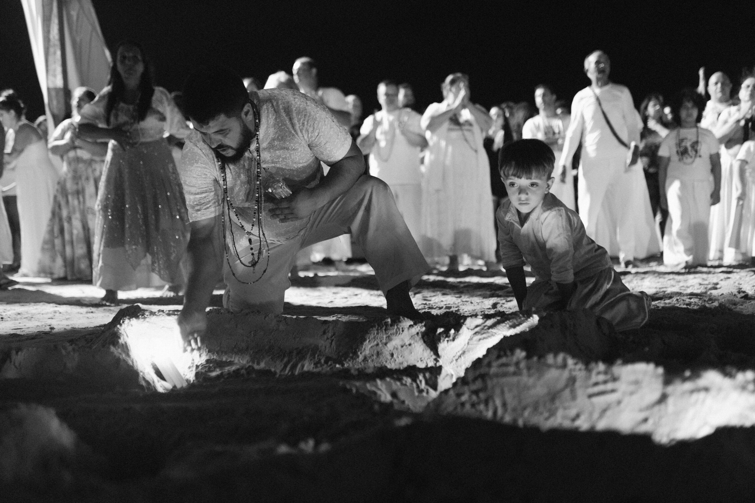 Galeria de Imagens. imagem 27: Fotografia em preto e branco, capturada em plano médio e ambiente noturno, retratando um momento de ritual coletivo sobre a areia da praia. Em primeiro plano, um homem adulto e um menino estão ajoelhados diante de uma vala escavada na areia, iluminada internamente por luzes intensas de velas, que projeta sombras dramáticas. O homem é branco, com barba e vestindo roupas claras rendadas e colares religiosos, está curvado, com uma das mãos estendidas dentro da vala, como se estivesse depositando ou organizando algo no interior iluminado. Ao lado dele, um menino branco, com expressão séria e concentrada — está ajoelhado e inclinado para frente, observando atentamente o ritual. Ele veste uma camisa clara de mangas compridas e calça do mesmo tom. Ao fundo, dezenas de pessoas observam a cena com respeito. A maioria está de pé, em silêncio, muitas com as mãos postas em sinal de reverência. Predominam roupas brancas ou claras, típicas de celebrações religiosas ligadas a tradições afro-brasileiras. A atmosfera geral da imagem é marcada pelo simbolismo do gesto, pela presença infantil e pela luz que emana do interior da terra — remetendo à ancestralidade, ao sagrado e à continuidade das tradições.