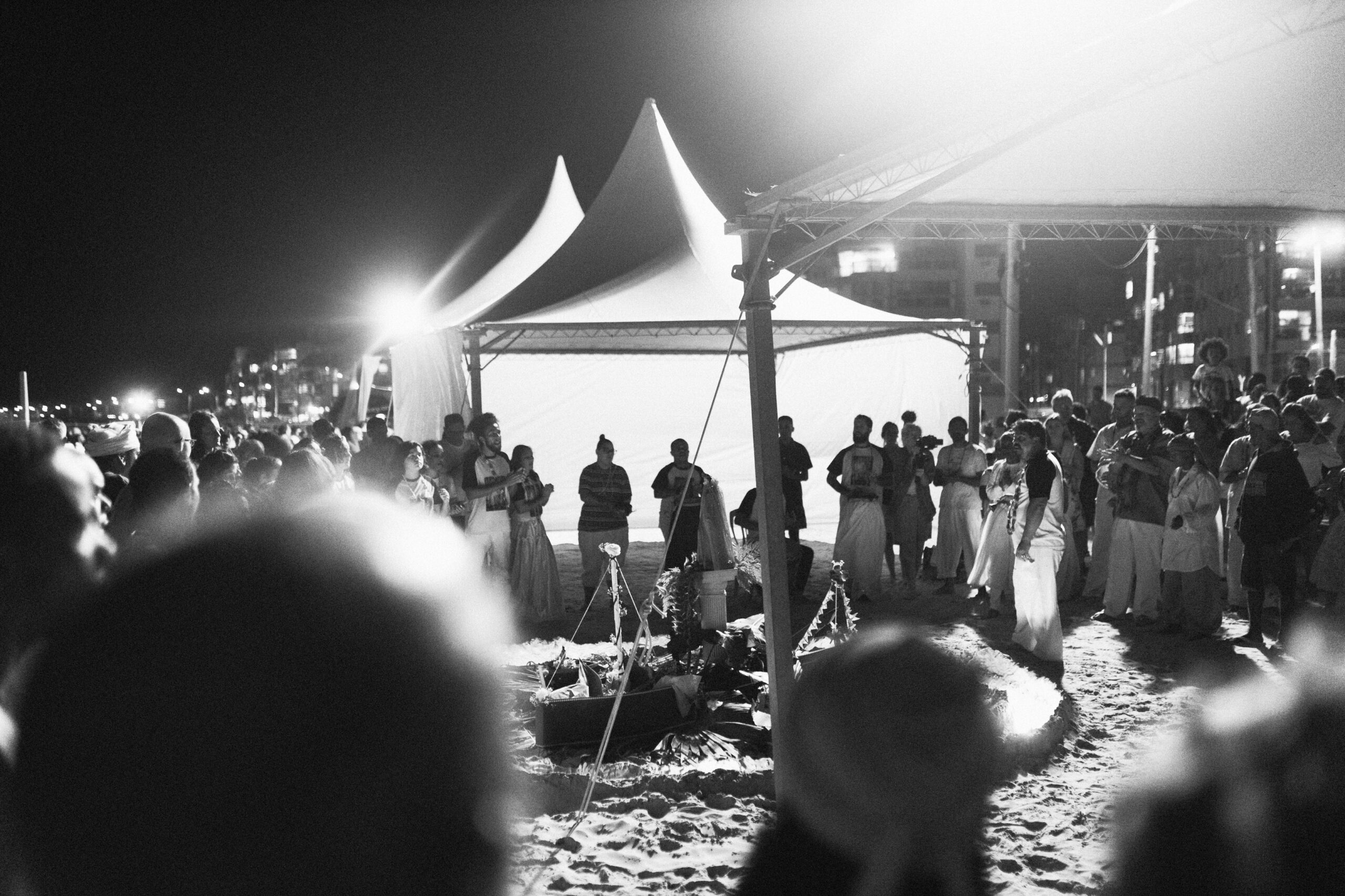 Galeria de Imagens. imagem 26: Fotografia em preto e branco, capturada em plano geral e ambiente noturno, retratando uma cerimônia religiosa coletiva na areia de uma praia. Em destaque, no centro da imagem, há uma estrutura ritualística cercada por cordinhas decoradas e flores, disposta sobre o chão de areia. Dentro da área delimitada, iluminada por uma luz intensa, há objetos simbólicos como: imagens, flores e velas. Ao redor da estrutura, uma multidão forma um grande círculo, observando em em atitude de respeito. Muitas pessoas vestem roupas brancas, trajes rendados ou panos tradicionais, demonstrando envolvimento religioso. A cena se passa sob grandes tendas brancas, montadas na areia, cuja iluminação forte contrasta com o fundo escuro da noite. À direita da imagem, vê-se parte da cidade ao fundo, com prédios iluminados.