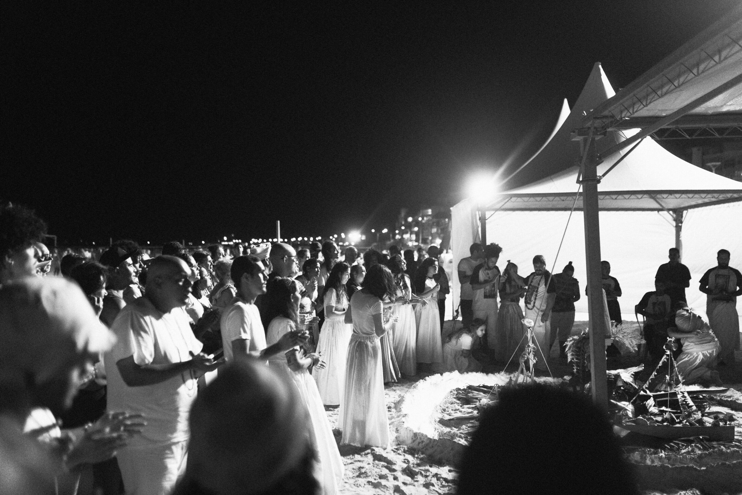 Galeria de Imagens. imagem 1: Fotografia em preto e branco, capturada em plano geral, mostrando uma cerimônia religiosa noturna realizada na areia da praia, sob tendas brancas iluminadas artificialmente. No centro da imagem, destaca-se uma estrutura ritualística sobre o chão de areia, composta por um círculo de elementos simbólicos, enfeites e objetos votivos - como estátuas, flores, folhagens de plantas, velas - demarcados com iluminação baixa e cordinhas decoradas. Um tripé com flores e uma pequena bandeira ocupa o centro da instalação. Ao redor da estrutura, uma multidão de pessoas observa. A maioria dos participantes veste roupas brancas ou claras — muitos em trajes longos ou com rendas —, tradicionais em celebrações religiosas afro-brasileiras. Algumas pessoas seguram flores ou objetos rituais nas mãos, outras estão com as mãos postas ou em gestos de reverência. À direita da imagem, uma tenda iluminada abriga mais pessoas, algumas sentadas, outras em pé, completando o cenário cerimonial. Ao fundo, vê-se a cidade iluminada e a linha da orla marítima com postes e prédios. O céu está escuro e limpo.