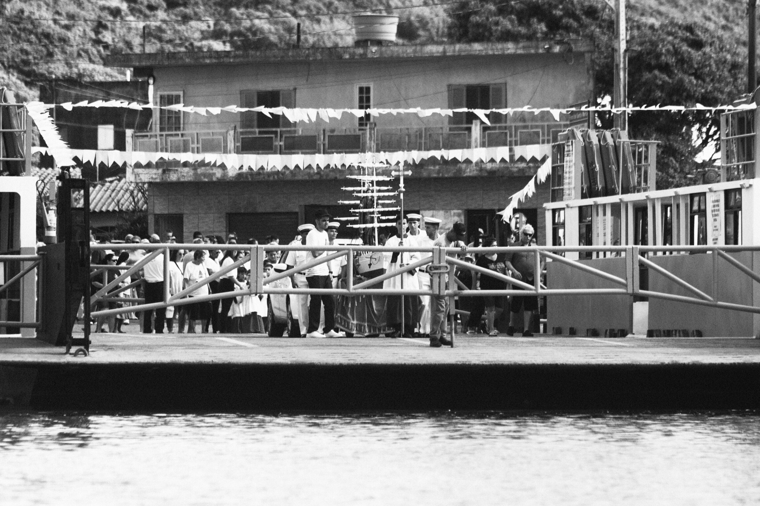 Imagem 4. Audiodescrição: Fotografia em preto e branco, capturada de frente para uma balsa atracada, vista da água. A balsa aparece parcialmente à direita da imagem, com sua rampa frontal ainda abaixada, conectando-se ao cais. O cenário mostra-se inserido em uma celebração religiosa popular, com bandeirolas brancas estendidas horizontalmente sobre a multidão, criando um clima festivo. Ao fundo, um grupo numeroso de pessoas aguarda, organizando-se em cortejo e no centro delas, está sendo conduzida uma estrutura simbólica de embarcação religiosa — com mastros e velas estilizadas — sobre a qual, embora parcialmente obstruída, é possível identificar a presença de uma imagem sacra. Militares da Marinha, com uniformes brancos e quepes, conduzem a plataforma cerimonial em direção à balsa. Crianças e adultos acompanham com atenção e reverência, alguns com trajes litúrgicos ou roupas brancas. A edificação ao fundo é simples, com fachada de alvenaria, janelas e varandas, típicas de comunidades litorâneas brasileiras. A imagem registra um momento de transição: o instante anterior ao embarque da santa na balsa, reforçando o simbolismo da proteção divina que atravessa as águas, elemento central em celebrações religiosas marítimas.