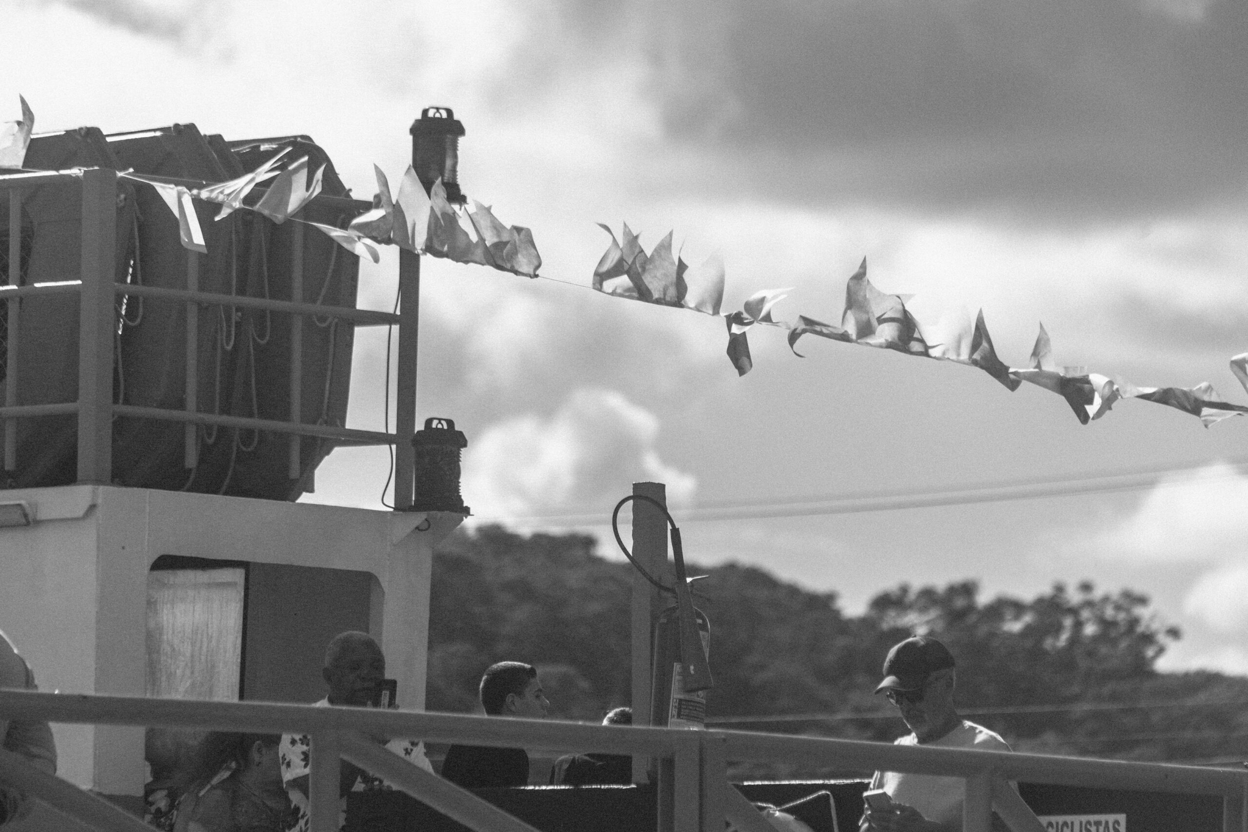 Imagem 12 da Galeria de Fotos. Audiodescrição: Fotografia em preto e branco, capturada à luz do dia, mostrando um detalhe da lateral de uma balsa fluvial ocupada por pessoas durante um momento de travessia ou festividade. No primeiro plano, há um corrimão metálico e, ao fundo, parte da estrutura da balsa — com paredes claras e elementos funcionais como grades e luzes de sinalização nos cantos superiores. Na parte superior da imagem, uma corda atravessa horizontalmente, ornada com bandeirolas pequenas e irregulares, agitadas pelo vento. No nível inferior, visíveis atrás do corrimão, estão várias pessoas. Um homem negro de cabelos curtos sorri discretamente enquanto olha para frente. À direita da imagem, outro homem — de pele clara, usando boné escuro e óculos, com a cabeça levemente inclinada. Ao redor deles, outras pessoas estão parcialmente visíveis, compondo um pequeno grupo. Ao fundo, fora de foco, pode-se ver a silhueta de árvores e vegetação baixa, além de linhas de energia cortando o céu. As nuvens estão espalhadas em um céu claro, sugerindo clima agradável. A imagem retrata um momento cotidiano e calmo, mas que, pela presença das bandeirolas e da concentração de pessoas, insinua um evento festivo ou religioso tradicional.