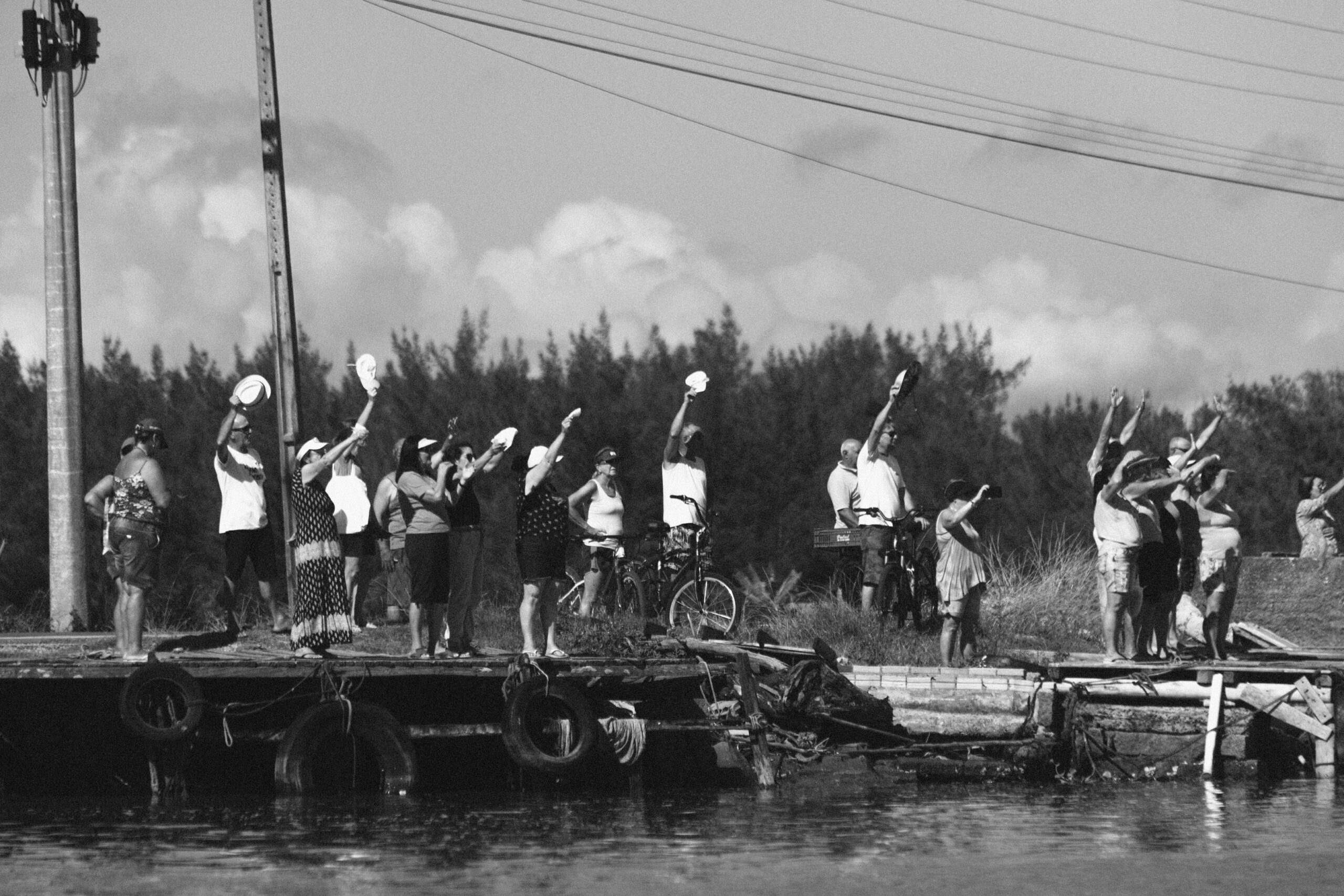 Imagem 13 da Galeria de Fotos. Audiodescrição: Fotografia em preto e branco capturada à luz do dia, mostrando um grupo de aproximadamente vinte pessoas reunidas sobre um cais rústico às margens de um corpo d’água. A maioria dos indivíduos está de pé, voltada para a direita da imagem, com os braços erguidos e acenando — alguns com chapéus, outros com panos brancos, em um gesto de despedida ou saudação festiva. O grupo é composto por homens e mulheres de diferentes idades, vestidos de maneira casual, com roupas leves típicas de clima quente. Algumas pessoas estão ao lado ou montadas em bicicletas, e nota-se ao fundo um trecho de vegetação densa com árvores baixas. Em primeiro plano, o cais exibe pneus pendurados nas bordas e estruturas desgastadas, características comuns de embarcadouros comunitários. O gesto coletivo e sincronizado do grupo, junto ao contexto visual das imagens anteriores, sugere que as pessoas estão despedindo-se possivelmente da balsa que conduz a imagem de Nossa Senhora dos Navegantes durante uma procissão fluvial. A cena transmite forte sentimento de reverência da comunidade local.