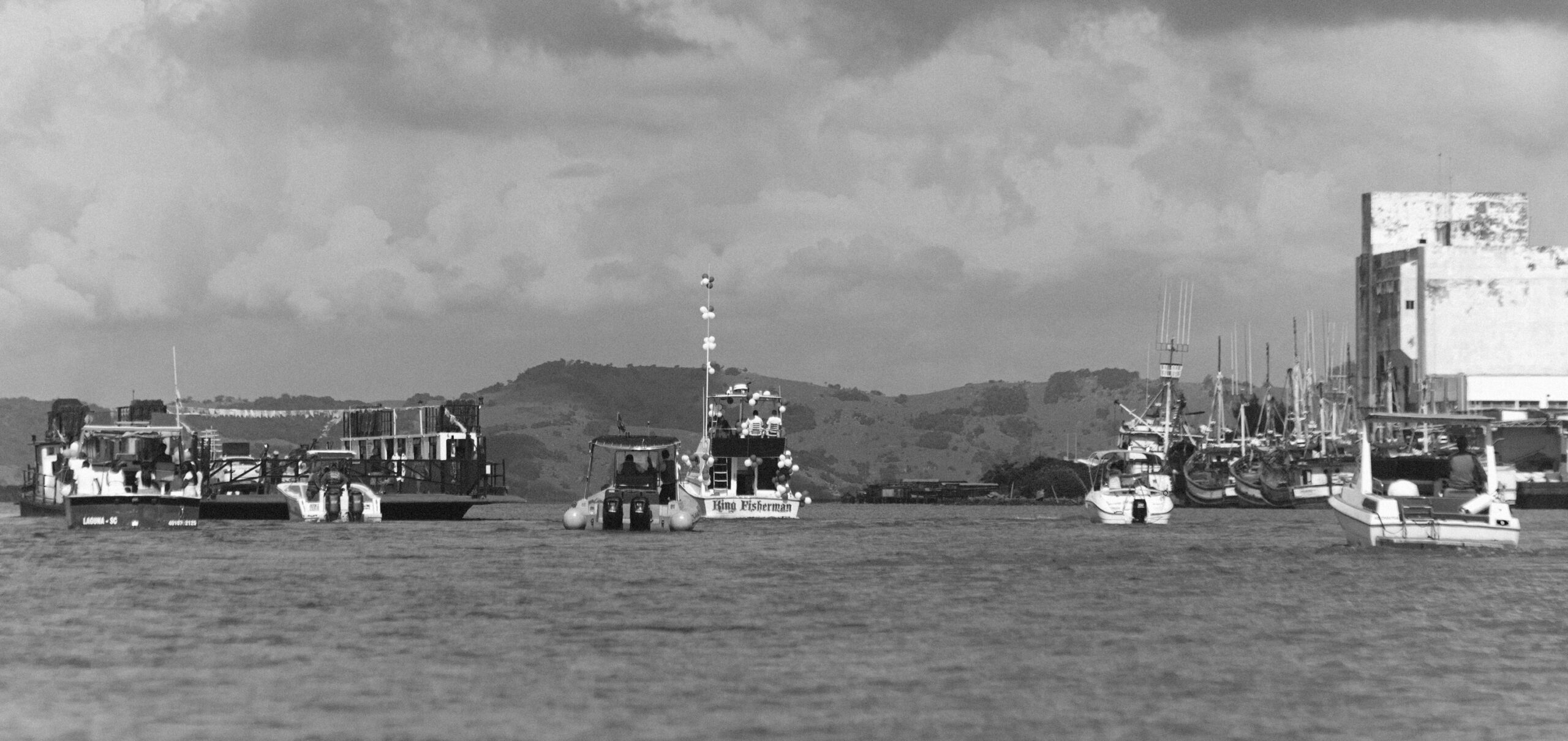 Imagem 15 da Galeria de Fotos. Audiodescrição: Fotografia em preto e branco, capturada durante o dia, com composição panorâmica de um trecho do mar com diversas embarcações. No plano central, observa-se uma barcaça larga, de estrutura robusta, provavelmente usada para transporte de veículos e pessoas. Atrás dela, vê-se uma série de outras embarcações menores e médias, incluindo barcos de pesca e lanchas, algumas enfeitadas com balões, como é o caso do barco ao centro nomeado King Fisherman. À direita da imagem, encontra-se um pequeno porto ou cais com vários barcos atracados lado a lado, junto a um grande edifício branco, de fachada desgastada, remetendo a um antigo armazém portuário. No fundo, observa-se uma paisagem de morro com vegetação densa, sob um céu encoberto por nuvens volumosas, que acrescentam dramaticidade à cena. O ambiente evoca uma procissão fluvial ou evento comemorativo náutico, reforçado pelos balões e pela quantidade de barcos alinhados. A água está calma e reflete suavemente a movimentação das embarcações.