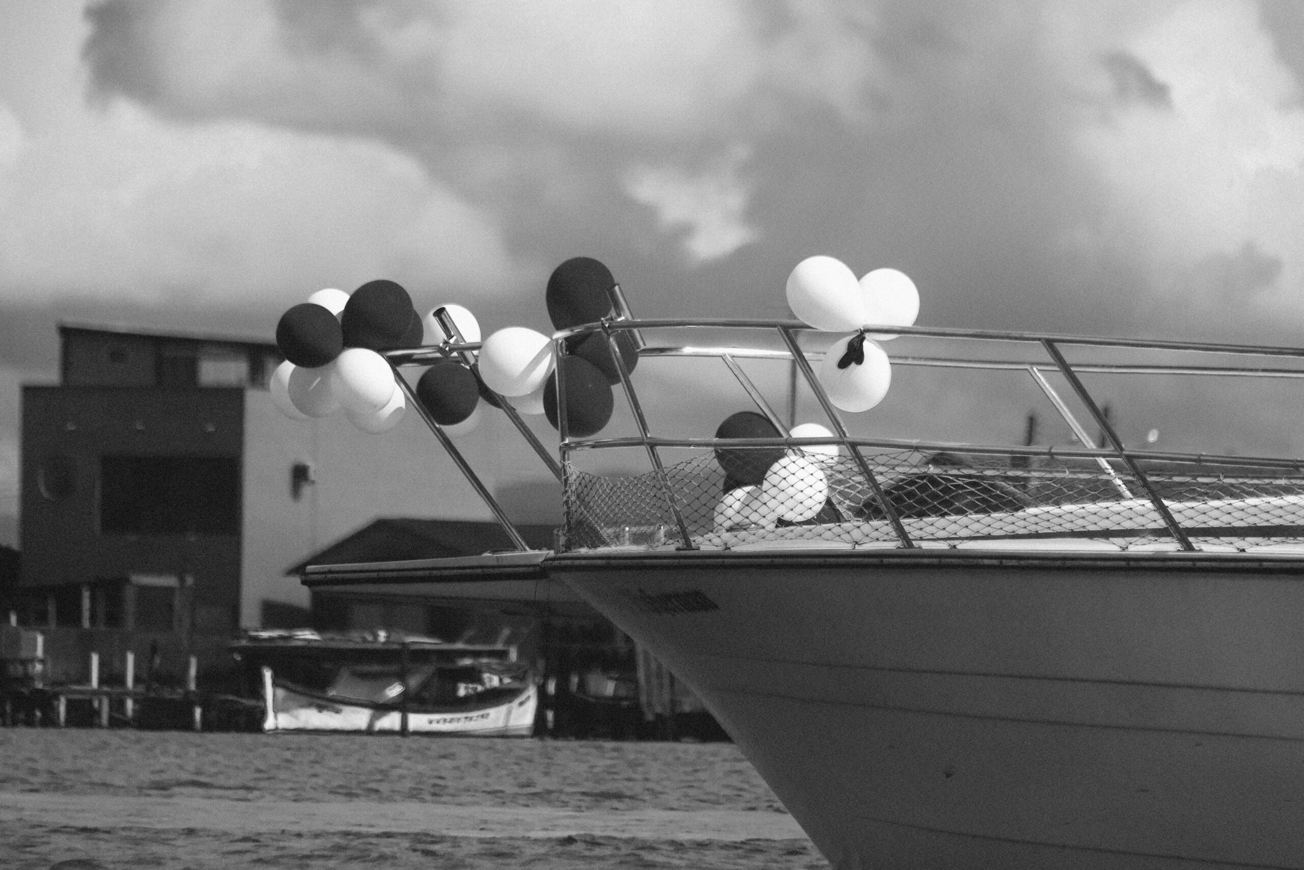 Imagem 17 da Galeria de Fotos. Audiodescrição: Fotografia em preto e branco retrata a proa de uma lancha decorada, apontando para a direita da imagem. A embarcação, com estrutura metálica e grades de proteção em malha de aço, está enfeitada com balões agrupados em pequenos arranjos. Os balões, em tonalidades claras e escuras (provavelmente coloridos no original), estão presos à grade da proa e ao suporte de metal inclinado. Destacam-se especialmente um par de balões brancos unidos ao centro da imagem, formando a aparência de um laço ou flor. O fundo mostra parte de um cenário urbano litorâneo. Ao longe, é possível ver casas e edificações baixas, incluindo um prédio de dois andares com fachada simples e janelas quadradas. Mais à esquerda, aparece parcialmente um barco de pesca atracado. O céu está tomado por nuvens densas e volumosas, criando uma atmosfera de luminosidade difusa e clima ameno.