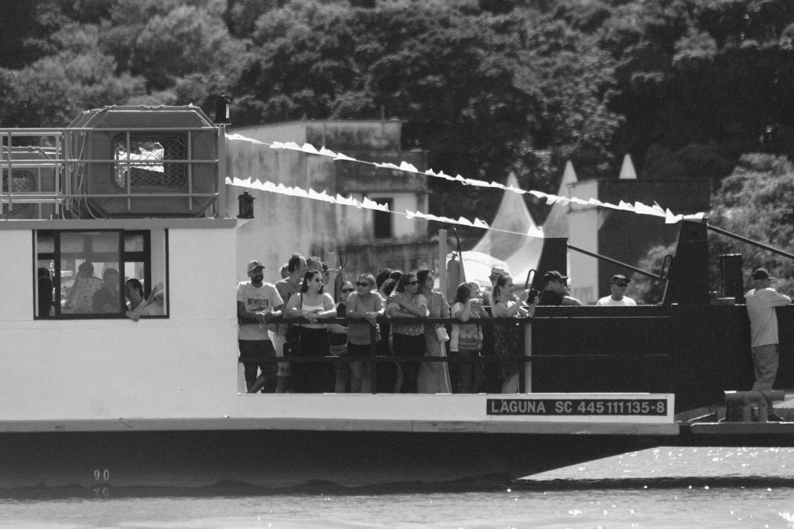 Imagem 19 da Galeria de Fotos. Audiodescrição: Fotografia em preto e branco mostrando uma balsa com diversas pessoas a bordo, navegando sobre águas calmas. A embarcação ocupa quase toda a largura da imagem. Na lateral da balsa, em destaque, há uma inscrição que identifica a embarcação: “LAGUNA SC 445111135-8”. A balsa está decorada com bandeirolas triangulares, estendidas em cordões diagonais acima da multidão, sugerindo um evento festivo. A bordo, há uma aglomeração de pessoas — homens, mulheres e crianças — que observam a paisagem ou conversam entre si. Alguns seguram celulares, enquanto outros apenas contemplam. Próximo à janela de uma cabine à esquerda, uma criança é visível olhando para fora, sentada no colo de um adulto. O fundo revela vegetação densa, com árvores em segundo plano, e uma construção simples ao centro, provavelmente um prédio antigo ou em ruínas. Também é possível ver as pontas de tendas triangulares brancas, sugerindo a presença de uma estrutura montada para evento na margem. A atmosfera geral da imagem é de celebração comunitária, com destaque para o aspecto humano da travessia e o uso coletivo do espaço fluvial.