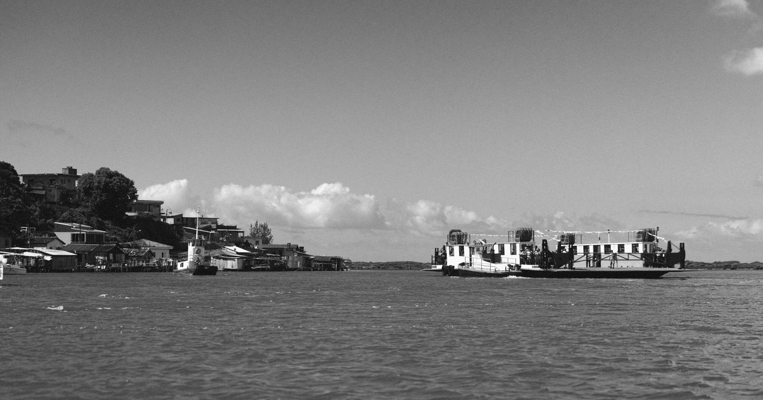 Imagem 20 da Galeria de Fotos. Audiodescrição: Fotografia em preto e branco de uma paisagem fluvial ampla, com uma balsa navegando da direita para o centro da imagem. A balsa é grande, de estrutura retangular e plana, com detalhes em branco e faixas escuras. Está enfeitada com bandeirolas triangulares ao longo da parte superior, sugerindo uma festividade. Algumas pessoas são visíveis a bordo, posicionadas próximas às bordas da embarcação. À esquerda da imagem, vê-se uma vila ribeirinha com casas simples e embarcações menores atracadas próximas a estruturas de madeira sobre o corpo d’agua. As edificações se estendem sobre uma encosta, em diferentes níveis, acompanhando o relevo da vegetação. No fundo, uma vegetação densa cobre o horizonte e o céu está parcialmente nublado, com nuvens esparsas contrastando contra o céu claro. A composição transmite uma sensação de tranquilidade, com destaque para a integração entre comunidade, natureza e navegação em um contexto cultural e cotidiano.