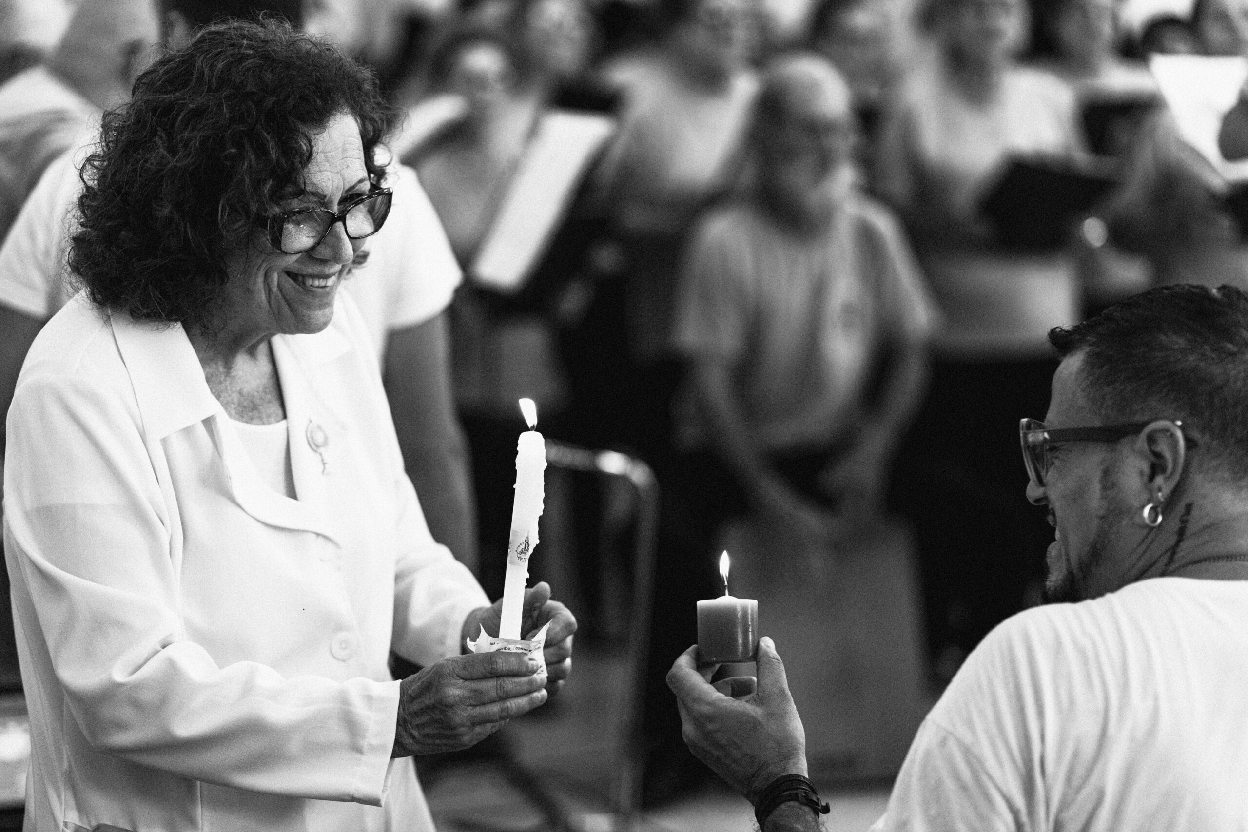 Imagem 21 da Galeria de Fotos. Audiodescrição: Fotografia em preto e branco, captada em ambiente fechado, retratando o momento simbólico de acendimento de uma vela. No lado esquerdo da imagem, uma mulher idosa de pele clara e cabelos cacheados escuros, com óculos grandes e expressão sorridente, veste um jaleco branco e segura uma vela longa e fina, já acesa. À sua frente, do lado direito, um homem branco de perfil, usando óculos, barba aparada e brinco circular, estende a mão com uma vela pequena e espessa, também acesa, em direção à vela da mulher, simbolizando um gesto de partilha. Ao fundo, o ambiente está preenchido por um grupo de pessoas que parecem participar de uma cerimônia ou celebração, com muitos segurando papéis ou livretos, possivelmente acompanhando um canto litúrgico ou ritual religioso. O foco da imagem está no gesto de acendimento e nas expressões gentis dos dois participantes em primeiro plano, transmitindo um clima de respeito, espiritualidade e comunhão.