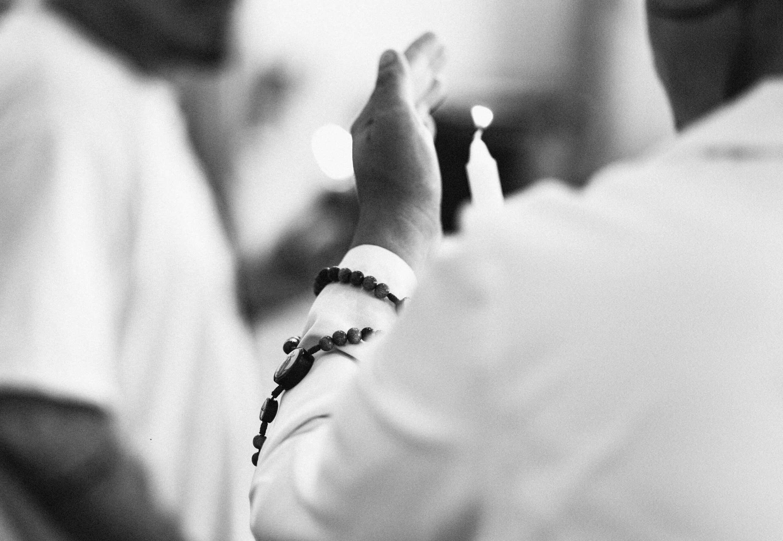 Imagem 5. Audiodescrição: Fotografia em preto e branco, capturada em plano fechado. A imagem mostra o braço erguido de uma pessoa com a palma da mão voltada para frente, como em um gesto de bênção, saudação ou oração. A pessoa veste uma camisa ou jaleco branco e usa um terço ou pulseira de contas escuras enrolada no punho, com cruz e medalha visíveis. Em segundo plano, levemente desfocado, vê-se a chama de uma vela acesa, sugerindo um ambiente cerimonial religioso. O restante da cena é intencionalmente desfocado, mantendo o foco no gesto simbólico da mão e nos elementos de fé representados pelo terço e pela vela. A iluminação suave e a composição intimista contribuem para a atmosfera de reverência, introspecção e espiritualidade.