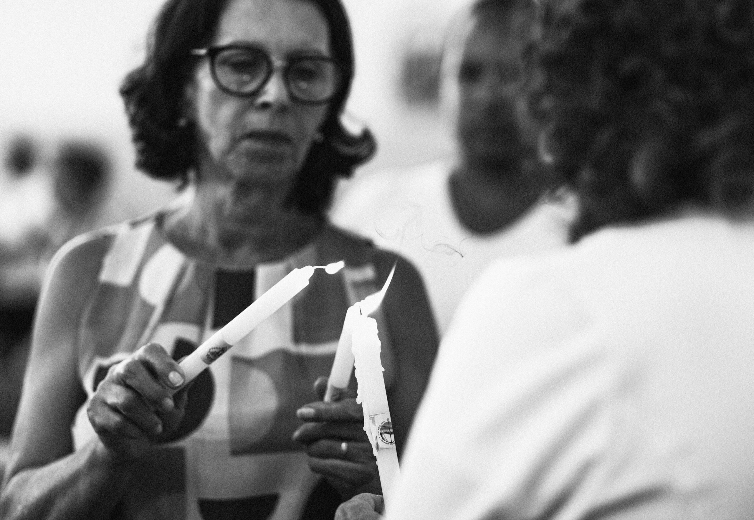 Imagem 23 da Galeria de Fotos. Audiodescrição: Fotografia em preto e branco, capturada em plano médio e foco seletivo. Em primeiro plano, à esquerda, uma mulher de pele clara, cabelos escuros e curtos, usa óculos de grau e está vestida com uma blusa sem mangas de estampa geométrica. Ela segura uma vela acesa com a mão direita, aproximando sua chama da vela de outra pessoa, fora de foco, à direita da imagem. Essa segunda pessoa, parcialmente visível de costas, veste uma roupa branca e também segura uma vela acesa, inclinada para receber ou compartilhar a chama. Entre as velas, é possível ver a pequena trilha de fumaça que se desprende da combustão, reforçando a atmosfera delicada do momento. O fundo da imagem está desfocado, revelando a presença de outras pessoas, sugerindo um ambiente coletivo e cerimonial religioso. A iluminação suave realça o caráter solene e íntimo da cena.