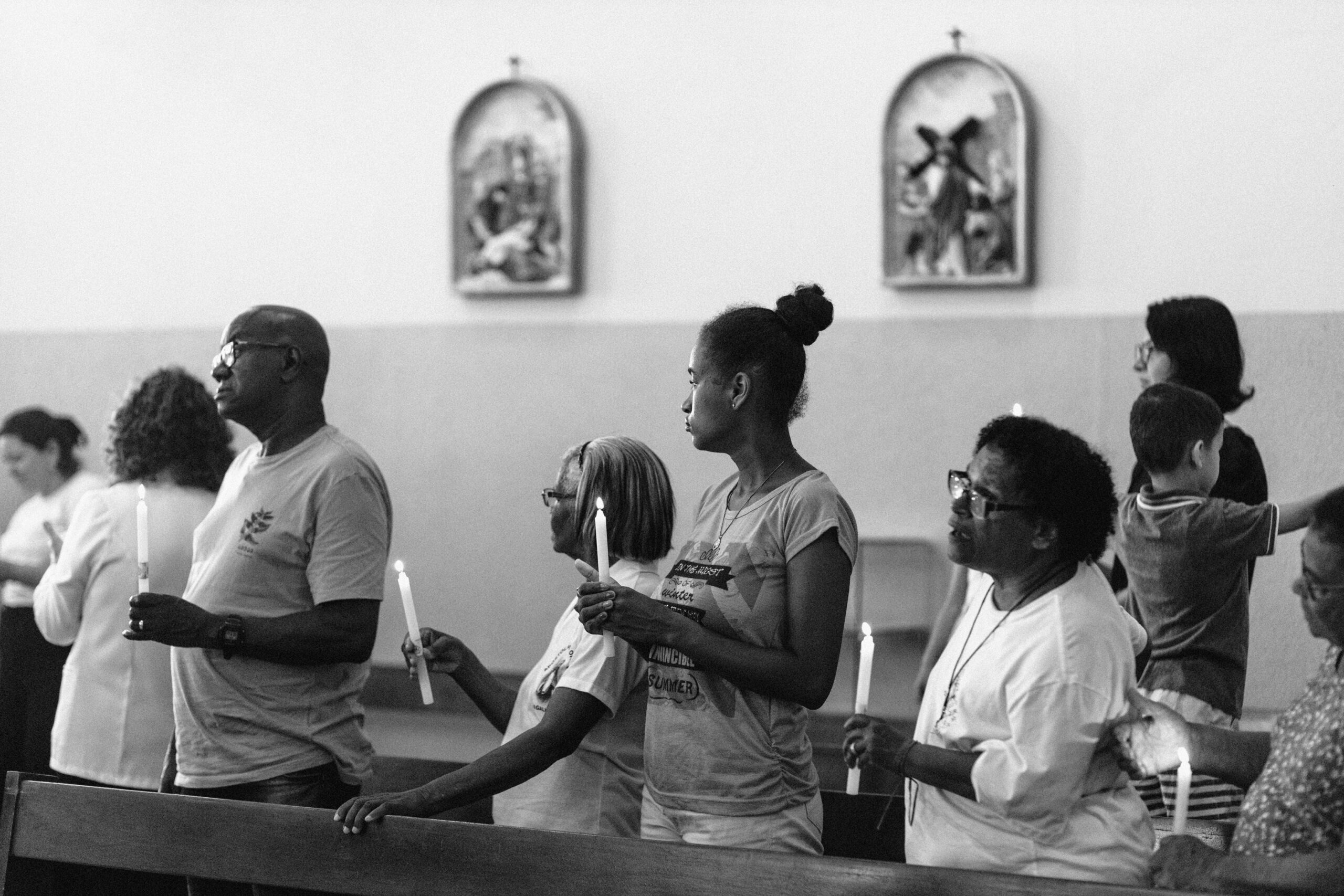 Imagem 7. Audiodescrição: Fotografia em preto e branco, feita no interior de uma igreja durante uma cerimônia religiosa. No primeiro plano, um grupo de pessoas está de pé entre os bancos, segurando velas acesas com expressão solene e respeitosa. São quatro pessoas negras no centro da imagem, todas adultas e aparentando idades variadas. À esquerda, um homem de cabeça raspada usa óculos e uma camiseta clara; ao seu lado, três mulheres — uma delas mais jovem, de cabelo preso em coque alto e camiseta com estampa; as outras duas com cabelos grisalhos, uma com camiseta branca e a outra com blusa clara e colar. Todos seguram as velas com firmeza, voltados para a esquerda da imagem, acompanhando o desenrolar do ritual. Ao fundo, desfocados, estão outras pessoas — incluindo uma criança — participando do mesmo momento litúrgico. Na parede da igreja, pendem dois quadros sacros em molduras ovais, com cenas religiosas. A cena transmite união, fé e espiritualidade, reforçada pelo simbolismo das velas acesas e a postura respeitosa dos participantes. A luz suave do ambiente e o preto e branco da foto acentuam o caráter introspectivo e devocional do instante registrado.
