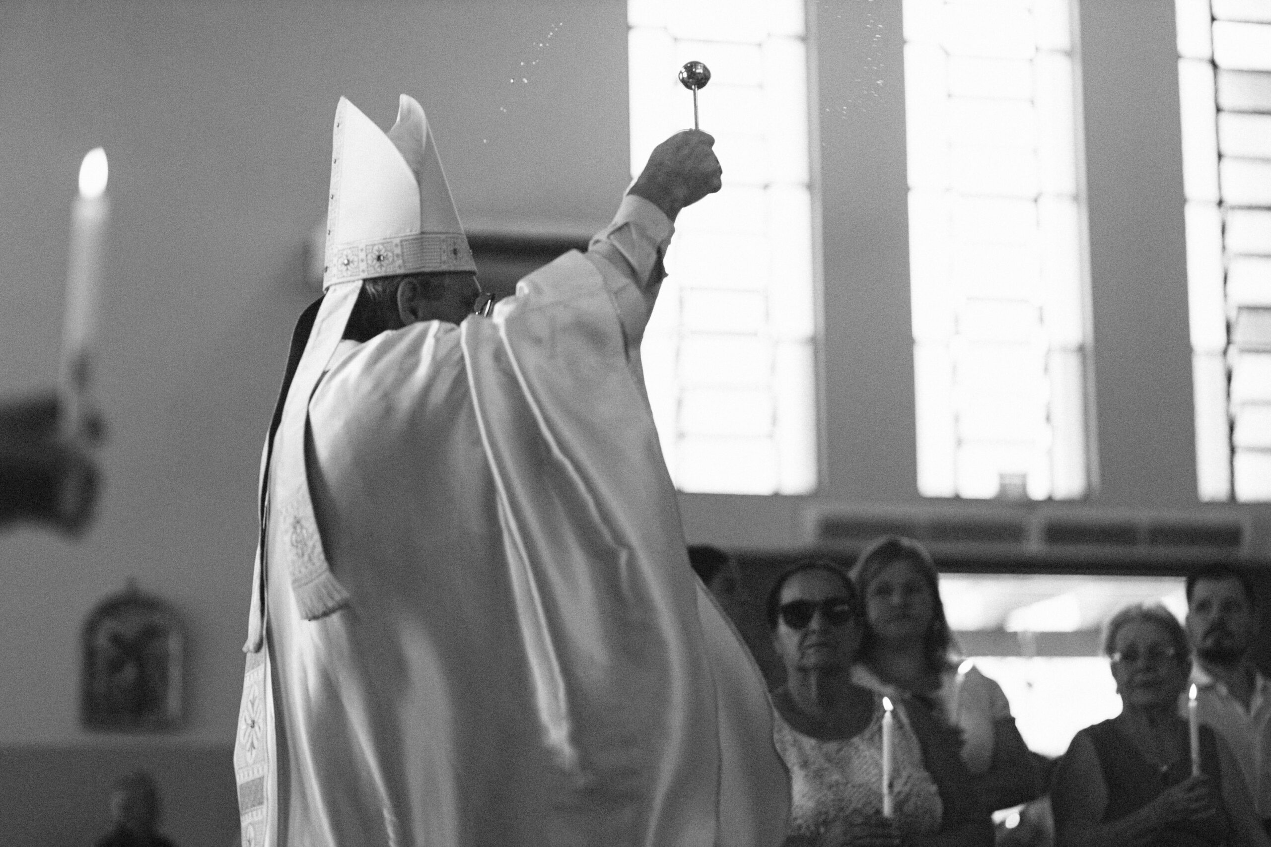 Imagem 26 da Galeria de Fotos. Audiodescrição: Fotografia em preto e branco capturada no interior de uma igreja durante um momento litúrgico. No centro da composição, um bispo é visto de costas, com destaque para a sua indumentária cerimonial. Ele veste uma longa veste litúrgica branca, com detalhes bordados, e usa um mitra ( tradicional chapéu alto e pontudo dos bispos ) decorado com uma faixa que cai pelas costas. O bispo está com o braço direito erguido, segurando um instrumento esférico na ponta de uma haste usado para aspergir água benta. Pequenas gotas d'água podem ser vistas suspensas no ar, indicando o instante exato da bênção. À frente, um grupo de fiéis, em sua maioria mulheres idosas, observa com reverência. Algumas seguram velas acesas. A luz suave entra pelas janelas altas e envidraçadas ao fundo, criando um ambiente etéreo e de devoção. À esquerda da imagem, fora de foco, há uma mão segurando uma vela. O clima da imagem é solene, simbólico e devocional, expressando um momento de fé coletiva e rito sagrado dentro do contexto cristão.