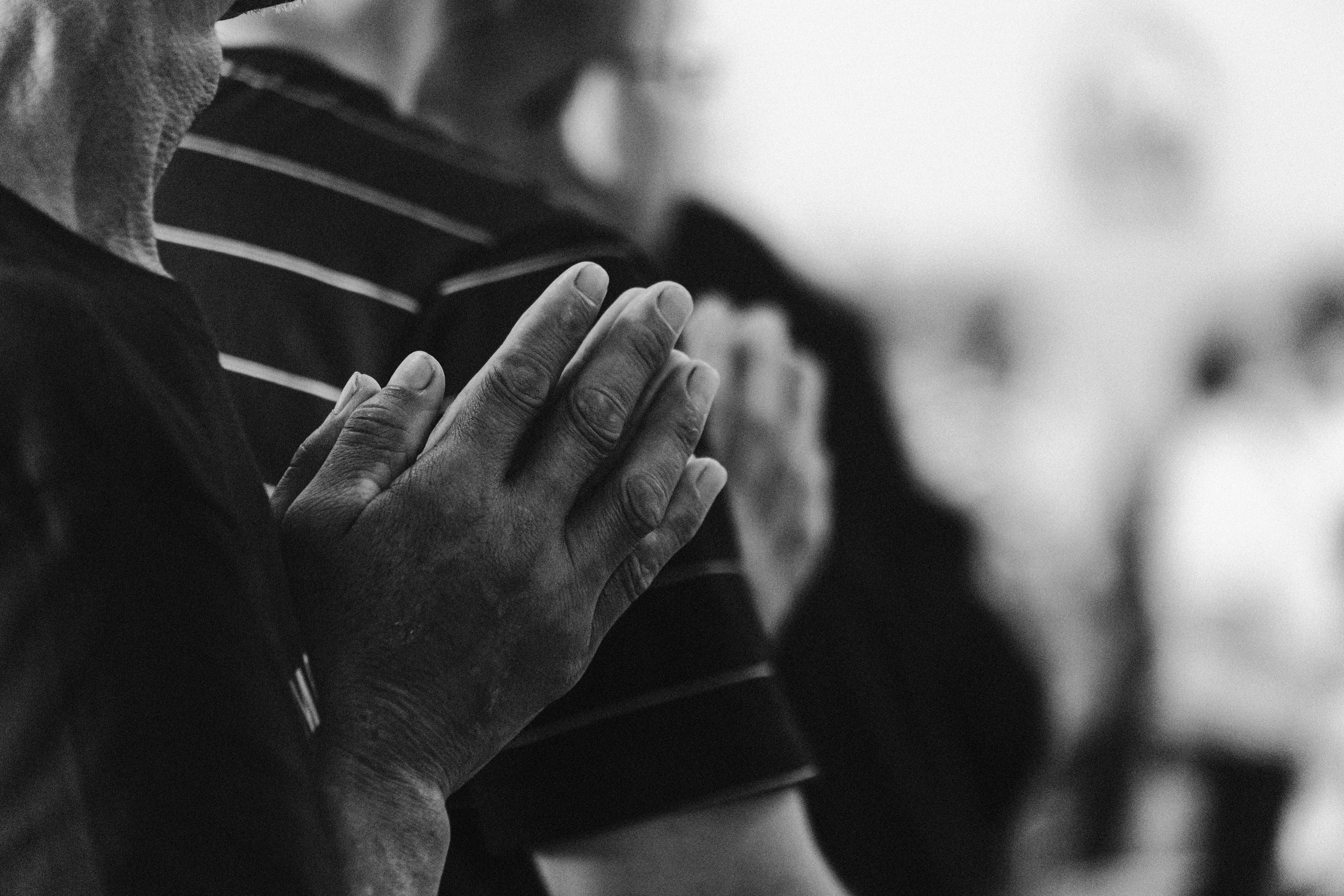 Audiodescrição da Imagem: Fotografia em preto e branco, em plano fechado, retrata mãos de uma pessoa branca idosa em posição de oração. As palmas estão unidas e os dedos apontam para cima, próximos ao peito, em gesto de reverência e concentração espiritual. As mãos apresentam textura visivelmente enrugada, indicando a passagem do tempo, e são parcialmente iluminadas, destacando seus contornos contra o fundo desfocado. Ao fundo, outras mãos também aparecem em posição semelhante, sugerindo que a cena ocorre em um ambiente coletivo de devoção numa celebração religiosa. As pessoas vestem roupas escuras, e uma delas usa camiseta com listras horizontais claras. A profundidade de campo é rasa, o que contribui para o foco intenso nas mãos em primeiro plano, enquanto o restante da cena permanece suavemente borrado. A imagem transmite introspecção, respeito e espiritualidade.
