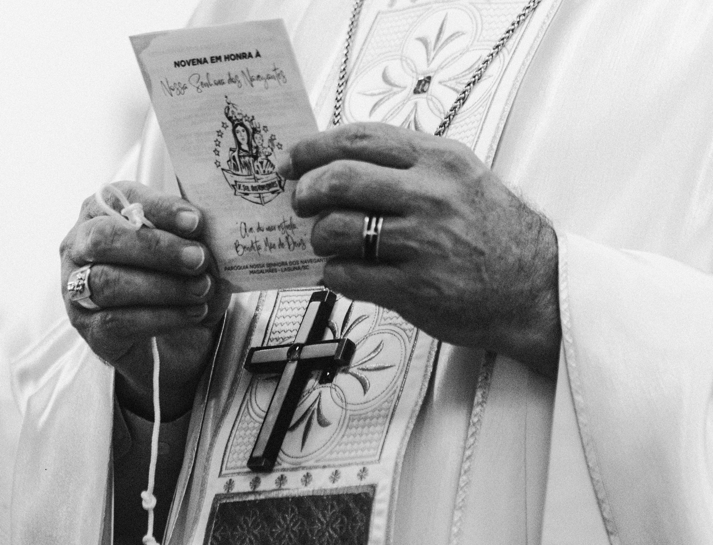 Imagem 29 da Galeria de Fotos. Audiodescrição: Imagem em preto e branco mostra em plano fechado as mãos de um sacerdote segurando um folheto de oração. O folheto, parcialmente visível, traz o título "Novena em honra à Nossa Senhora dos Navegantes", com uma ilustração da santa com o Menino Jesus nos braços, ambos cercados por estrelas e posicionados sobre um barco estilizado. Abaixo, há uma frase manuscrita: "Clara do mar estrelado, protegei-nos ó Mãe de Deus". Também é possível ler o nome da "Paróquia Nossa Senhora dos Navegantes de Magalhães – Laguna/SC". As mãos masculinas, de uma pessoa branca, firmes e adornadas com dois anéis — um em cada mão — sustentam o papel. Na mão esquerda, nota-se ainda um cordão branco enrolado nos dedos, possivelmente parte de um rosário. O traje litúrgico é claro, com bordado floral visível no paramento e uma cruz preta pendurada no peito, presa a um cordão grosso e escuro. A imagem transmite solenidade, reverência e espiritualidade, reforçada pelo vestuário litúrgico e pelo foco exclusivo nas mãos e nos elementos simbólicos da fé cristã.
